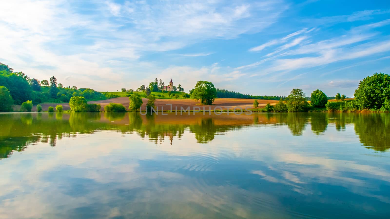 Romantic landscape with small church on the hill reflected in the pond. Sunny summer day with blue sky and white clouds. St. Peter and Pauls church at Bysicky near Lazne Belohrad, Czech Republic.