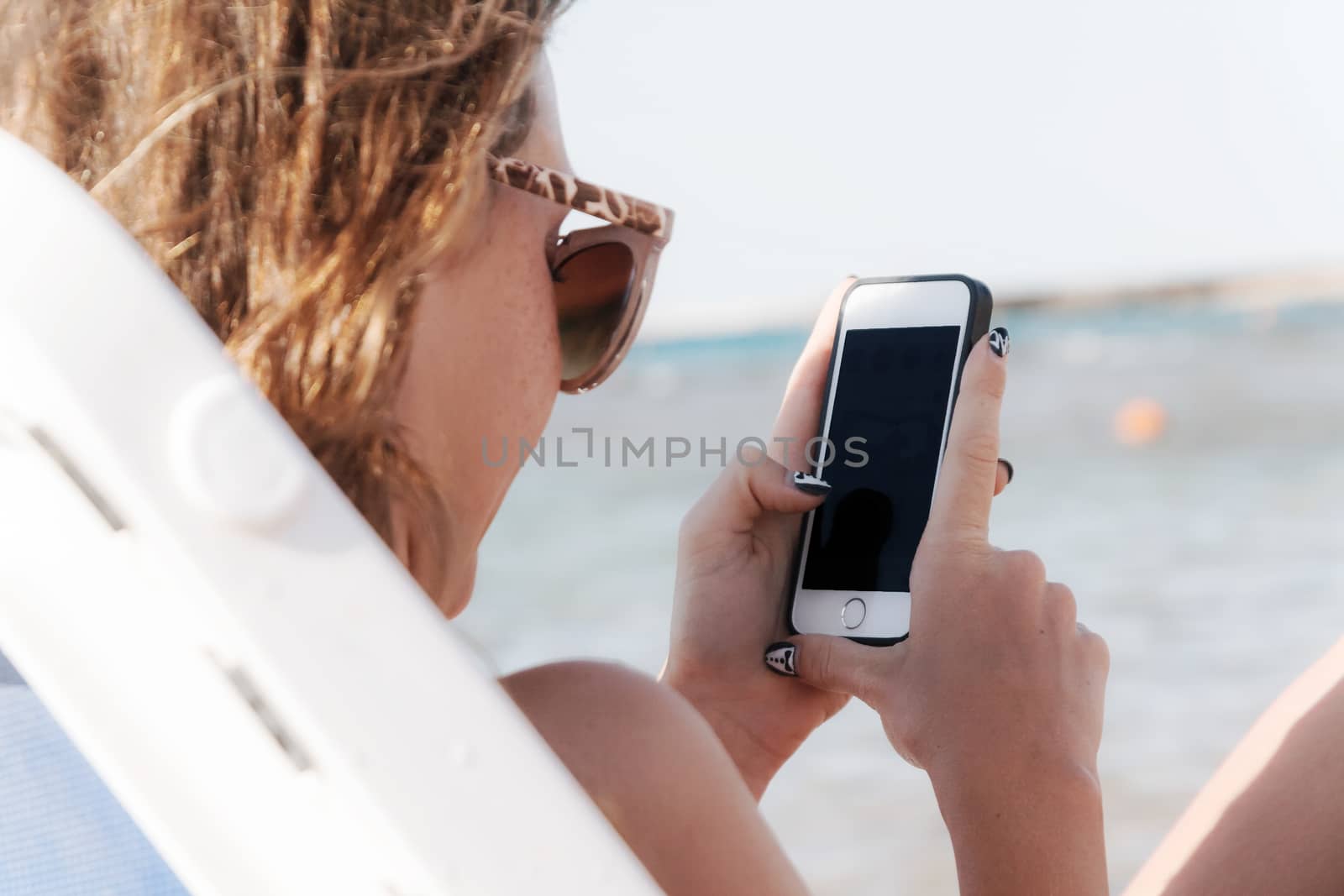 A young tanned woman looks into a smartphone through sunglasses  by Tanacha