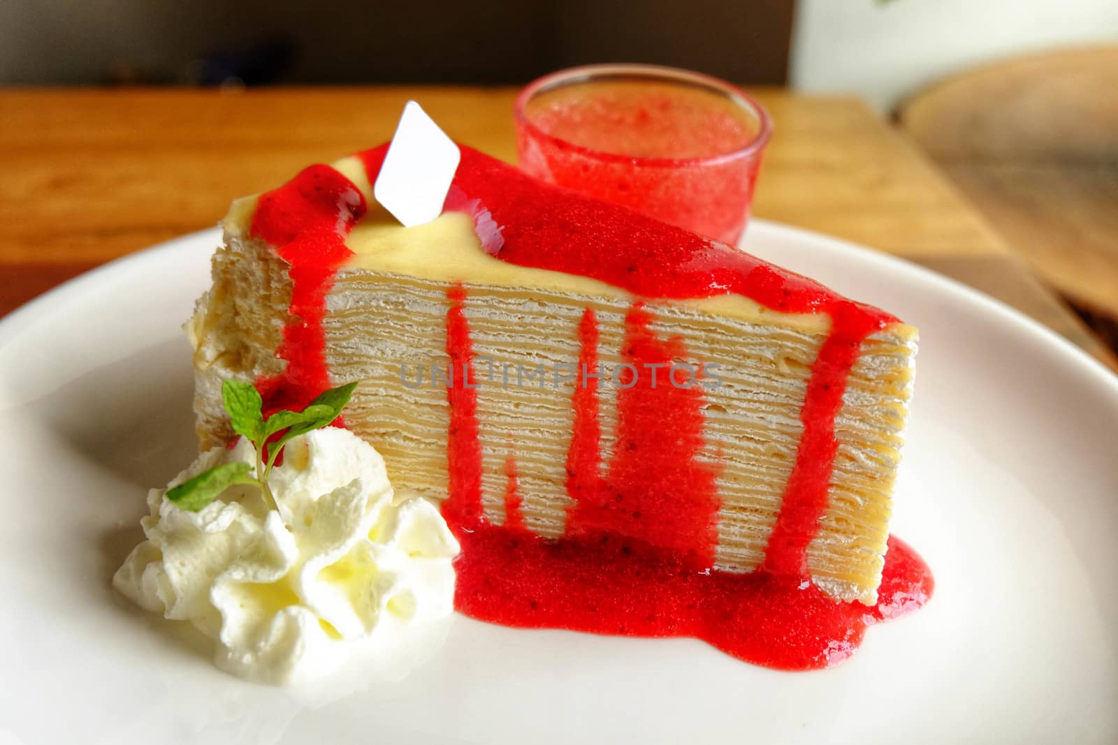 Crepe cake with cream and strawberry on top and sauce in ceramic cup on white plate.