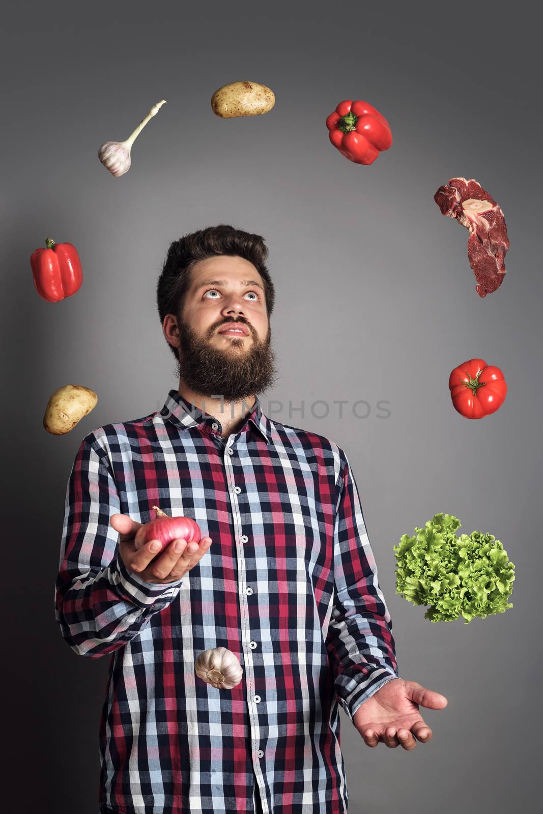 Cooking man concept, smiling bearded man in checked shirt, juggle meat and vegetables, studio shot on gray background