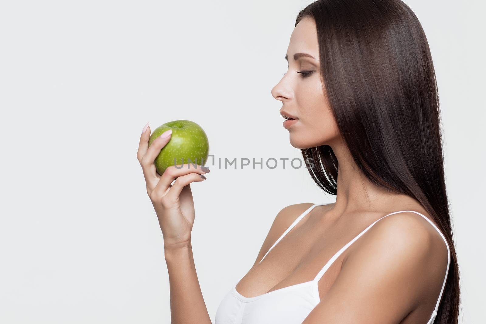 Side view of attractive brunette in sports bra biting green apple while standing on white background.