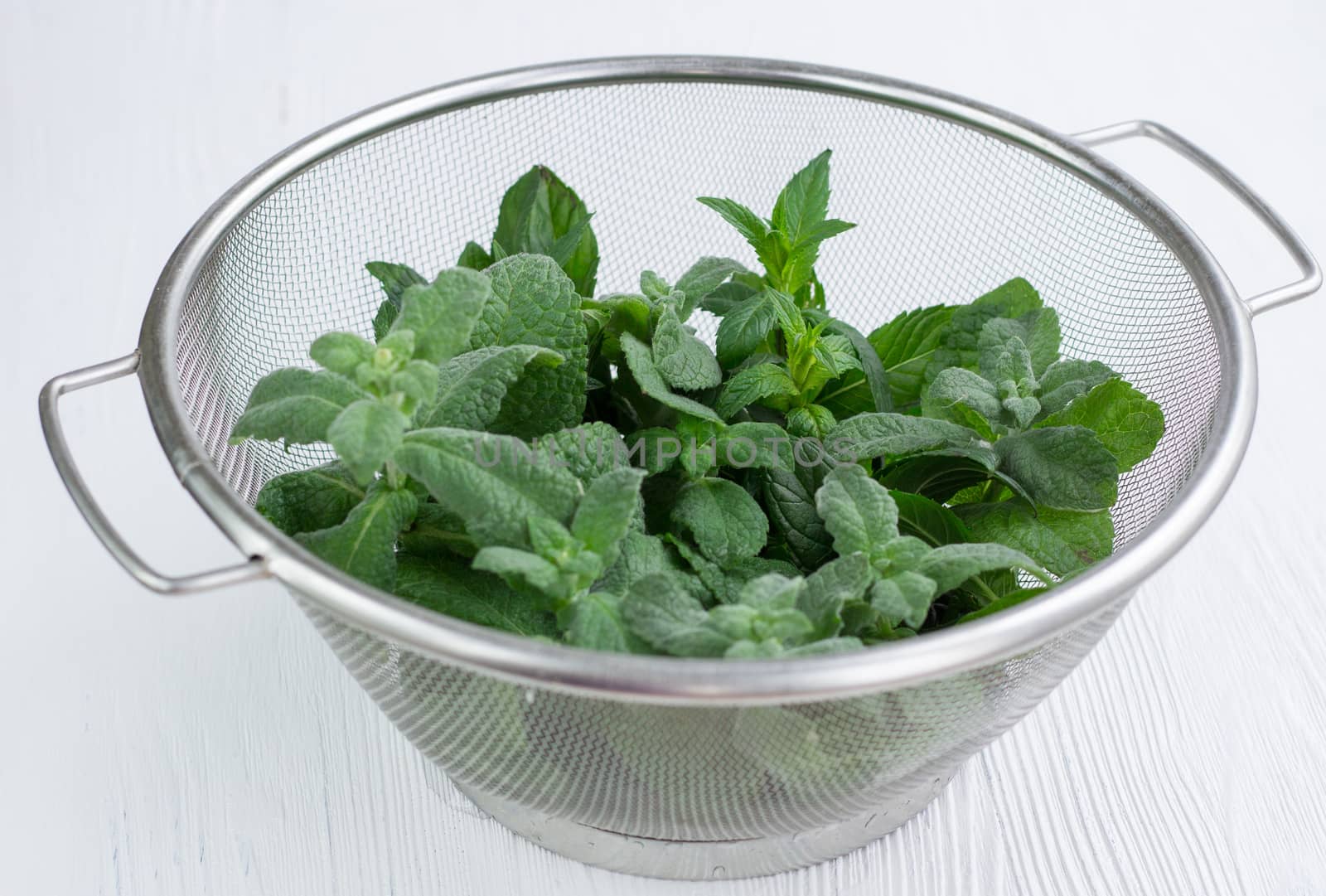 Green herbal mix of fresh mint and melissa herbs in stainless metal strainer bowl on white wooden background