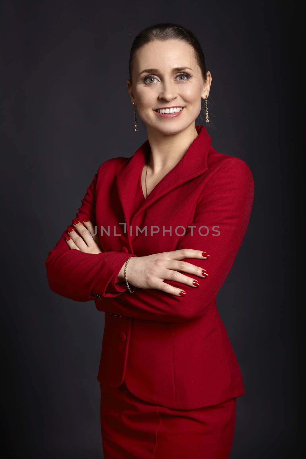 Portrait of elegant young business woman in red skirt suit with white smile and crossed arms, on black background.