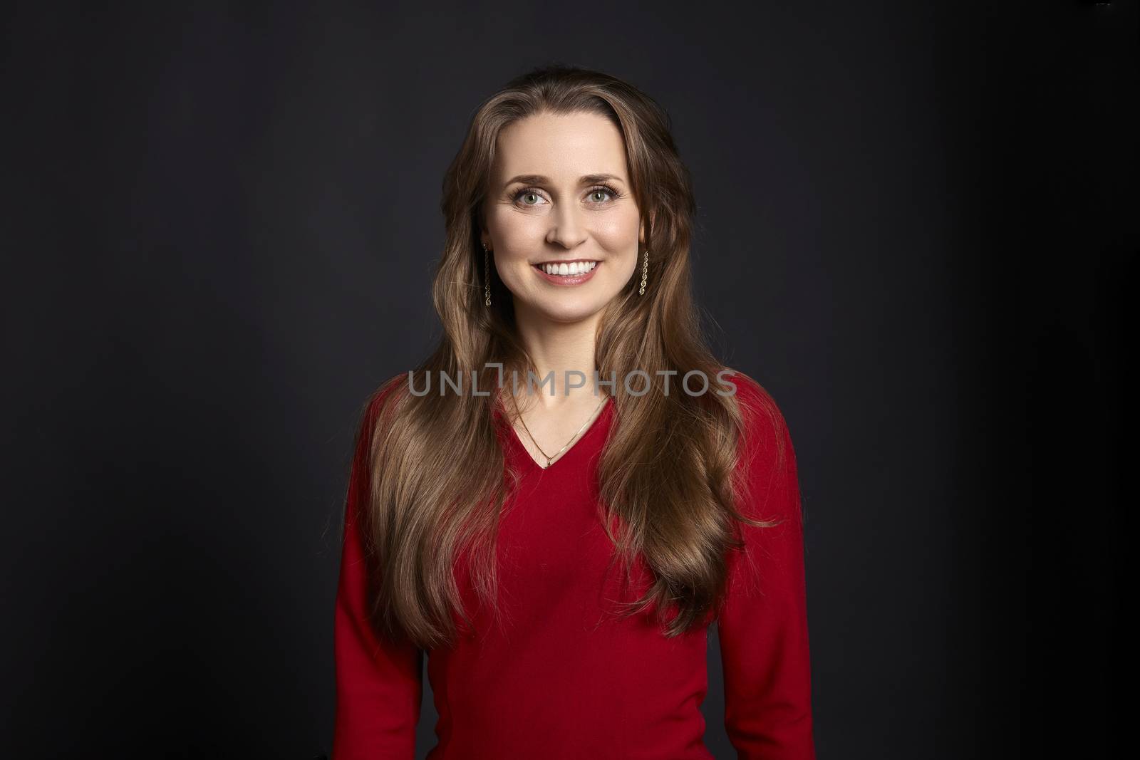 Studio portrait of young smiling woman in red dress with long hair, white smile and green eyes on black background.