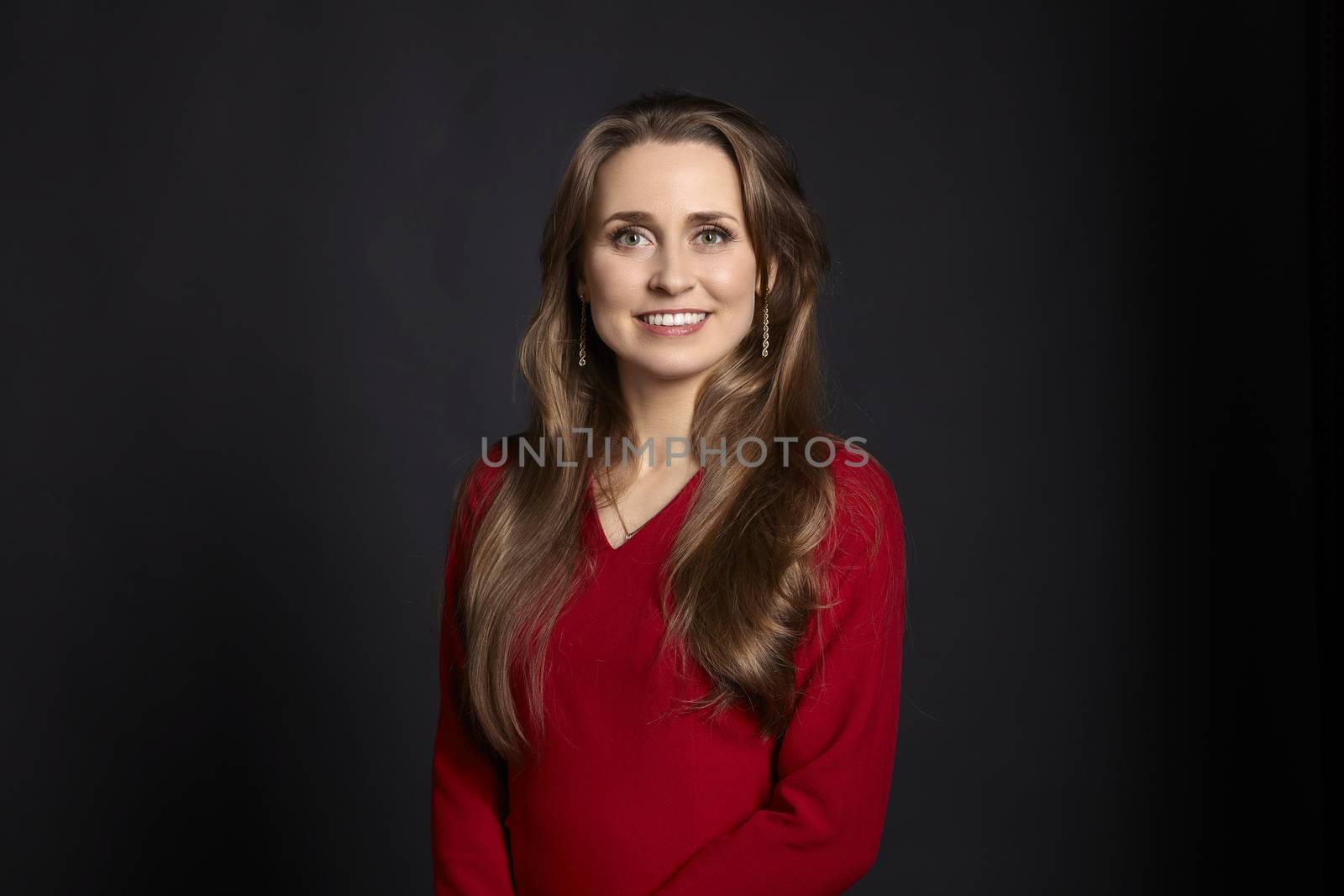 Studio portrait of young smiling woman in red dress with long hair, white smile and green eyes on black background.