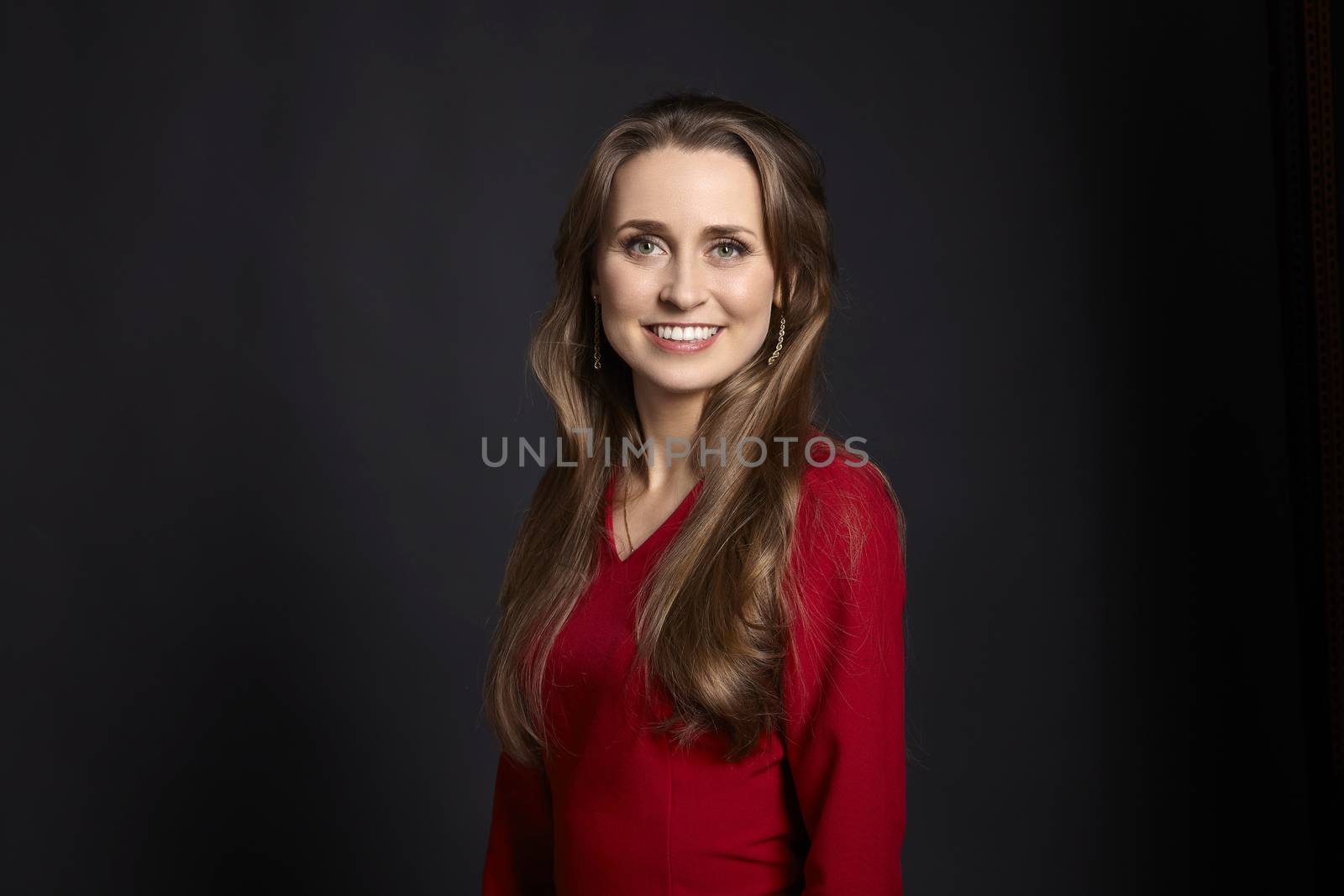 Studio portrait of young smiling woman in red dress with long hair, white smile and green eyes on black background.