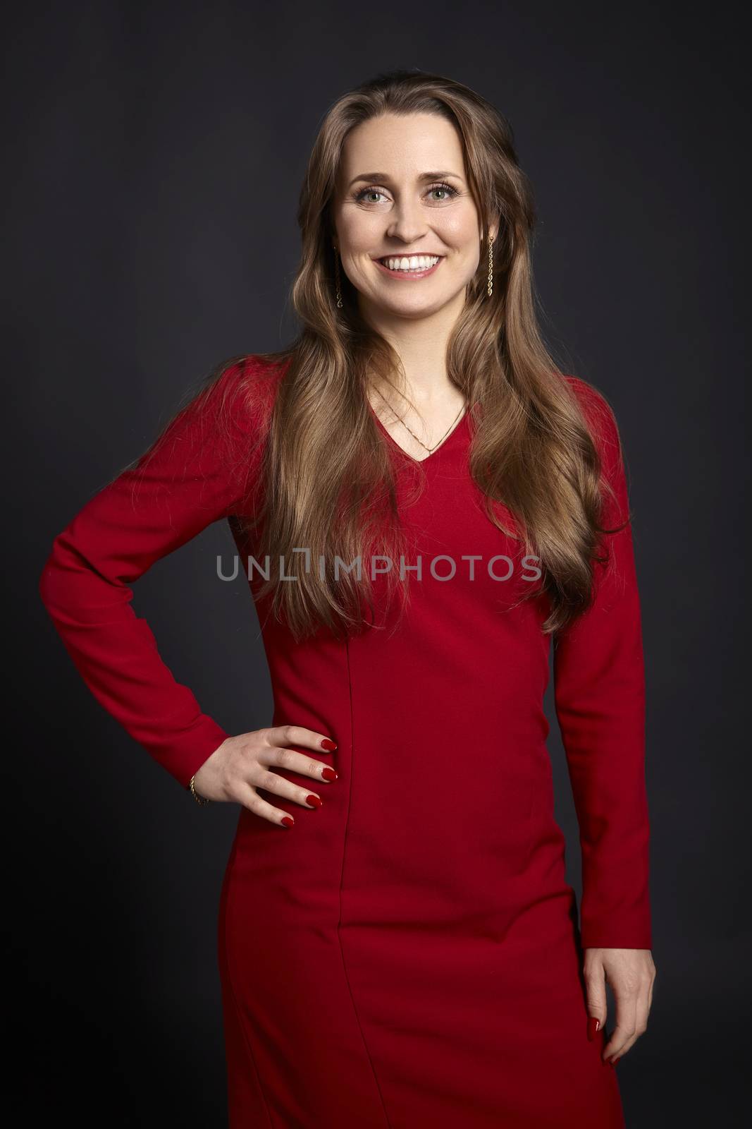 Studio portrait of young cheerful woman with long hair, white smile, green eyes and hand on hip on black background.