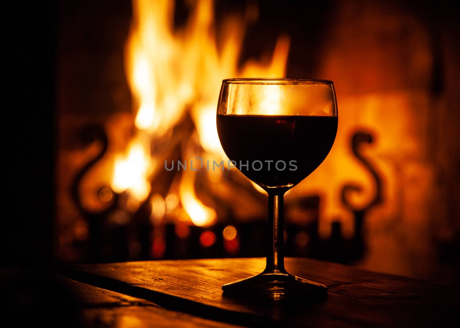 Glass of red wine on the wooden table with burning fire on the background. Evening relax on cozy place. Dark medieval style winery.