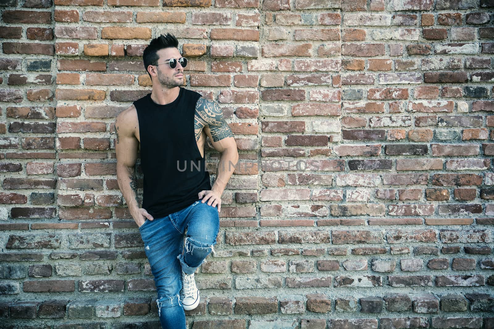 Attractive muscular man standing outdoor leaning against old brick wall, looking to a side