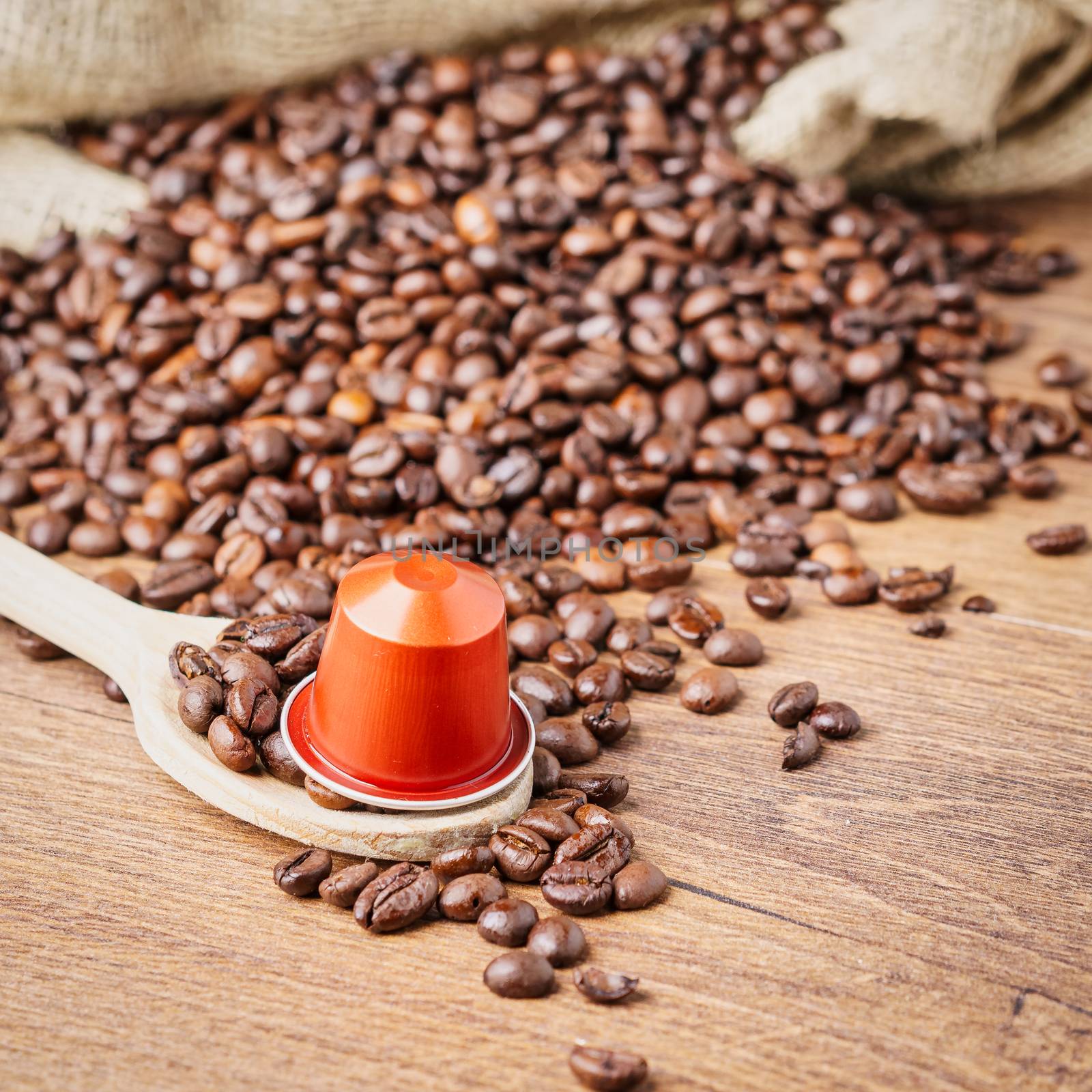 In the foreground a coffee capsule on wooden spoon and   roasted coffee beans with burlap sack on blur wooden background,top view.