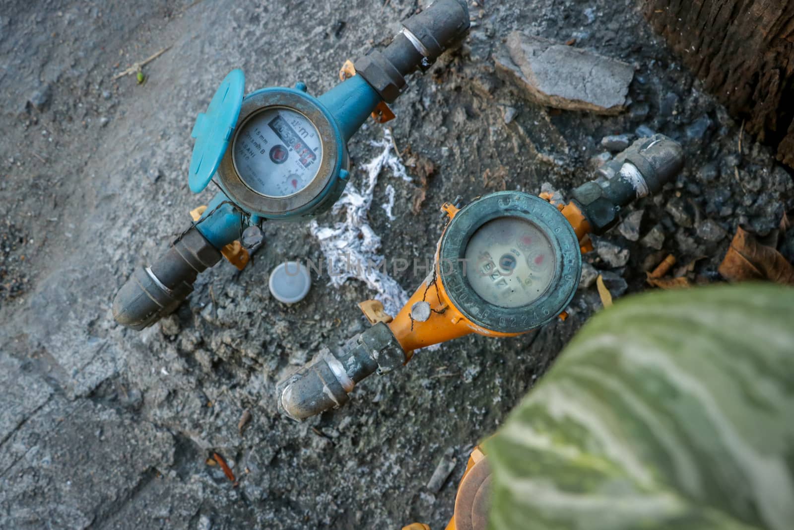 Vintage Orange and Blue Water Meters on Dirty Concrete/ Stone Texture in the Garden with Green Leaves in Foreground. Street Photography Bangkok Thailand.