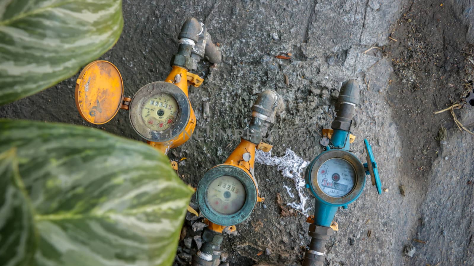 Vintage Orange and Blue Water Meters on Dirty Concrete/ Stone Texture in the Garden with Green Leaves in Foreground. Street Photography Bangkok Thailand.