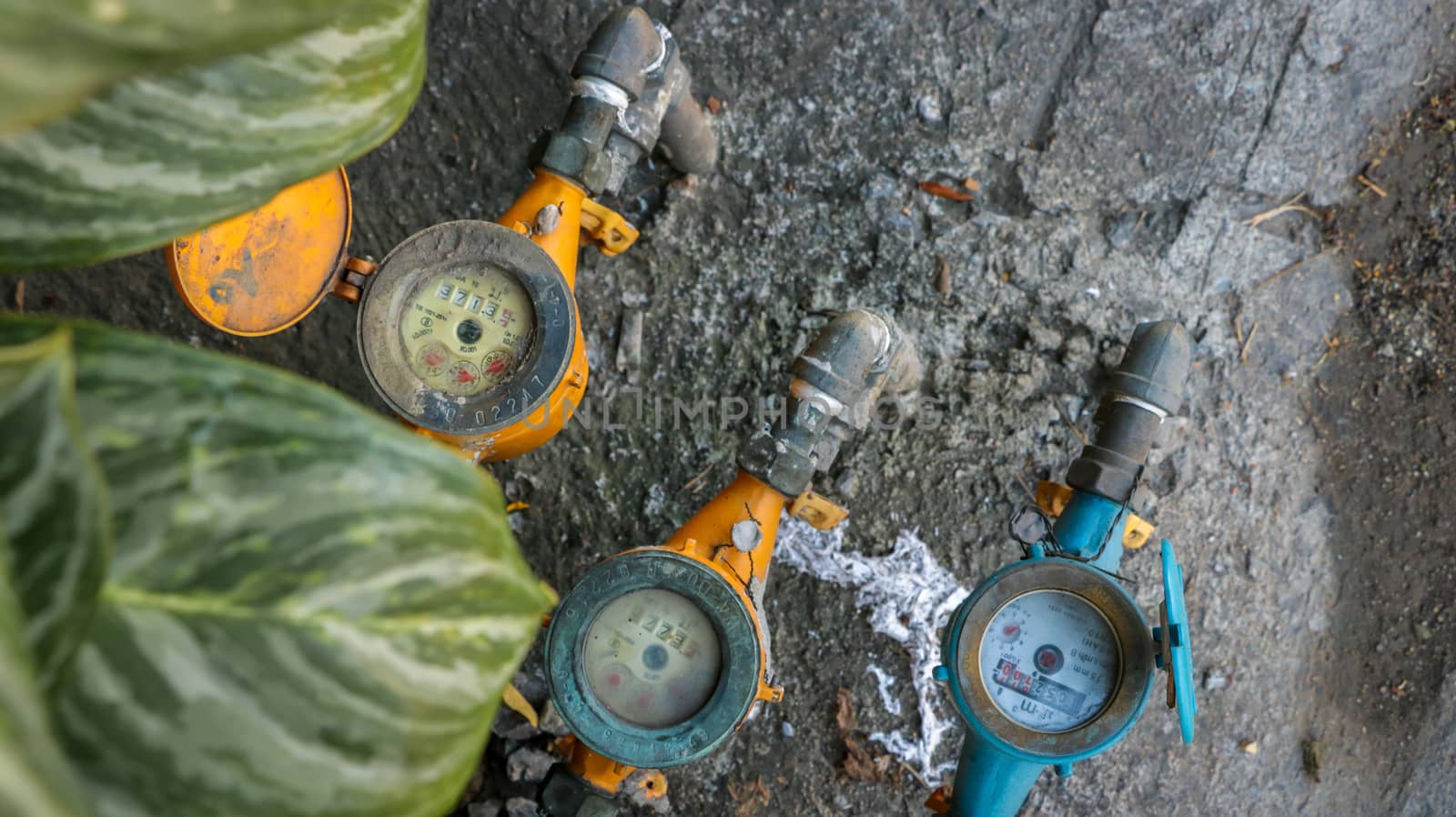 Vintage Orange and Blue Water Meters on Dirty Concrete/ Stone Texture in the Garden with Green Leaves in Foreground. Street Photography Bangkok Thailand.