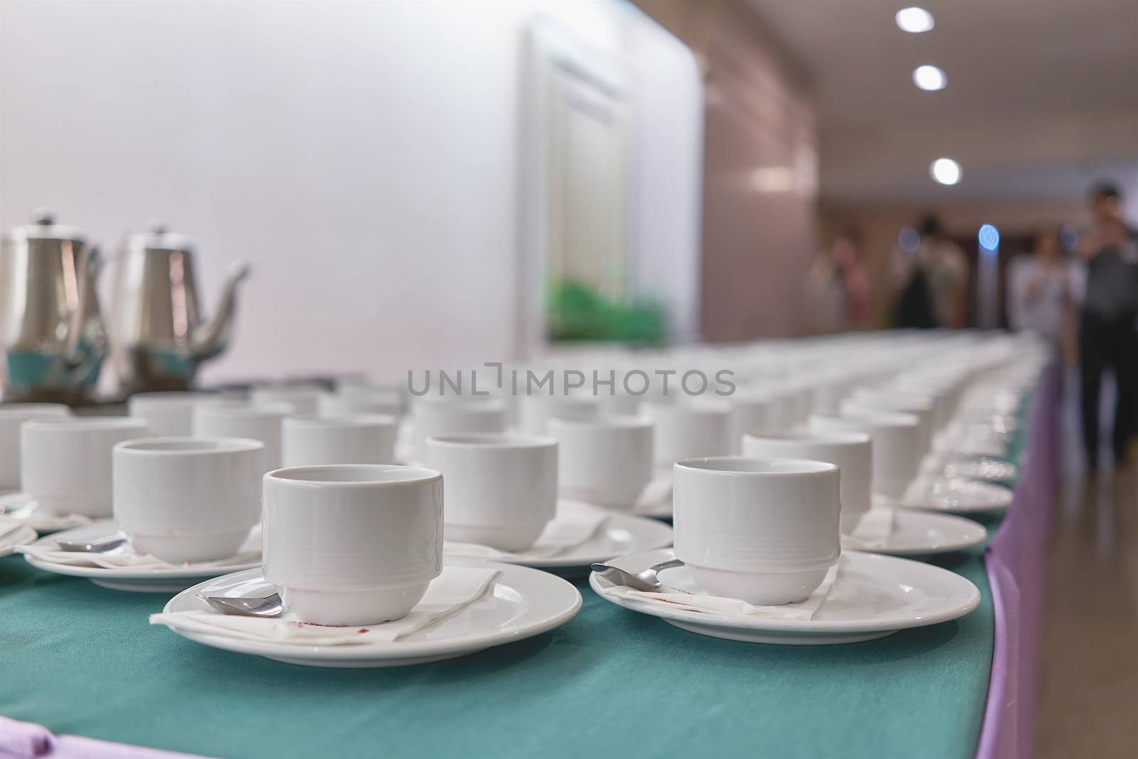 Empty white cups of coffee with spoon and tissue on saucer are placed in a row on green and purple table have blur coffee pots and people as background at conference in the hotel.