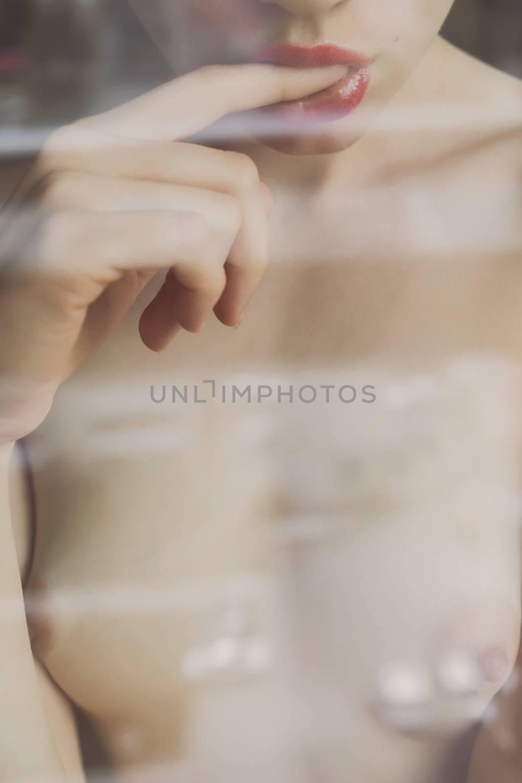 Crop view through glass of faceless naked young woman sexually putting finger in mouth