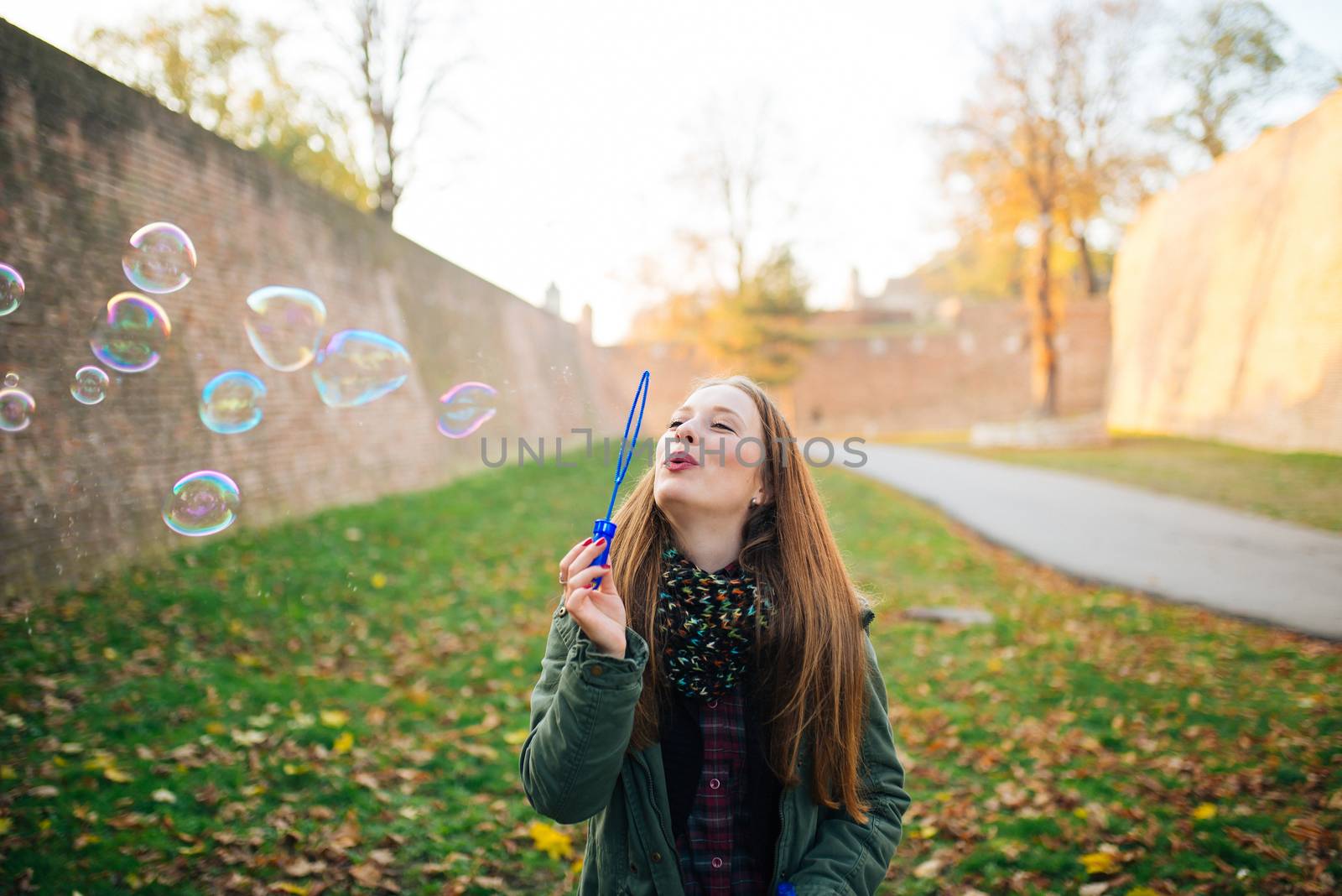 Attractive young woman is enjoying making a bubbles in the public park. Ground covered with autumn leaves.