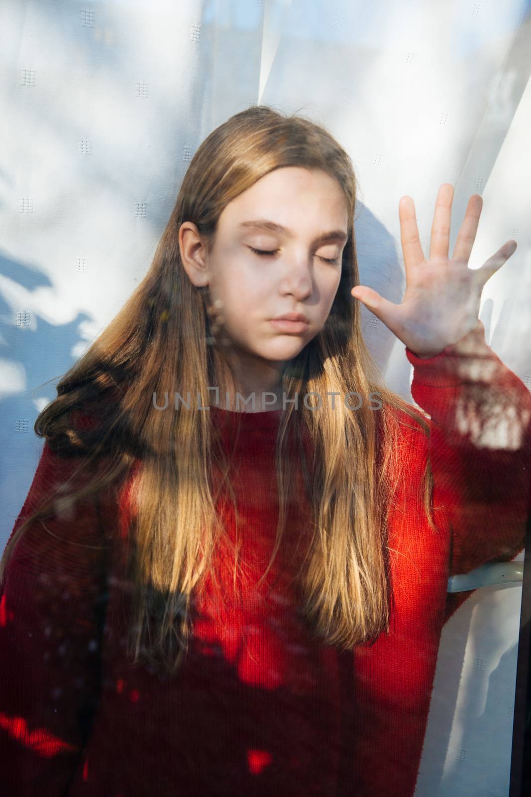 Sad cute little girl suffering depression and standing behind the window during a rainy day with raised hand 