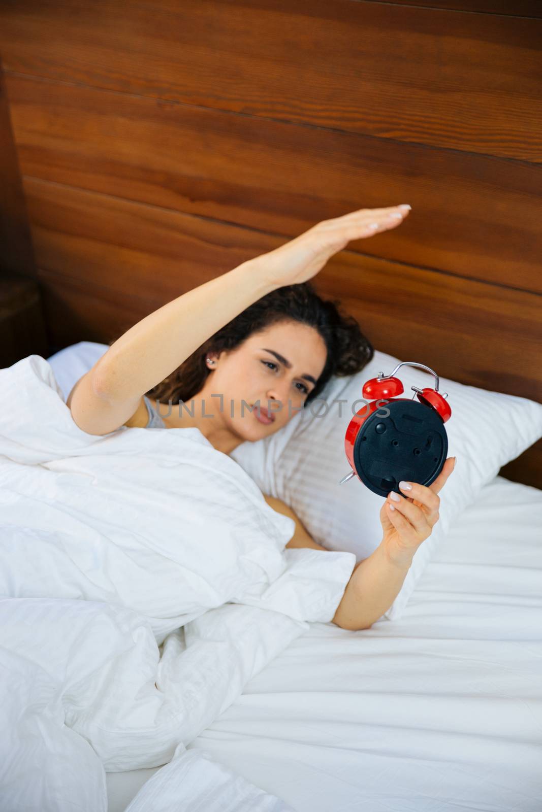 Beautiful woman awakened by alarm clock in the bed at morning time. A woman is unhappy from noisy sound of alarm clock and trying to turn it off. 