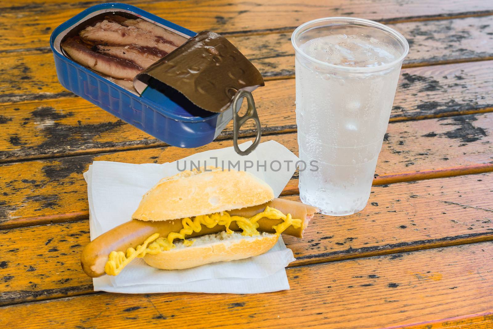 Bockwurst in bun, a can of fish, mug with mineral water on a brown wooden table on a white napkin.
