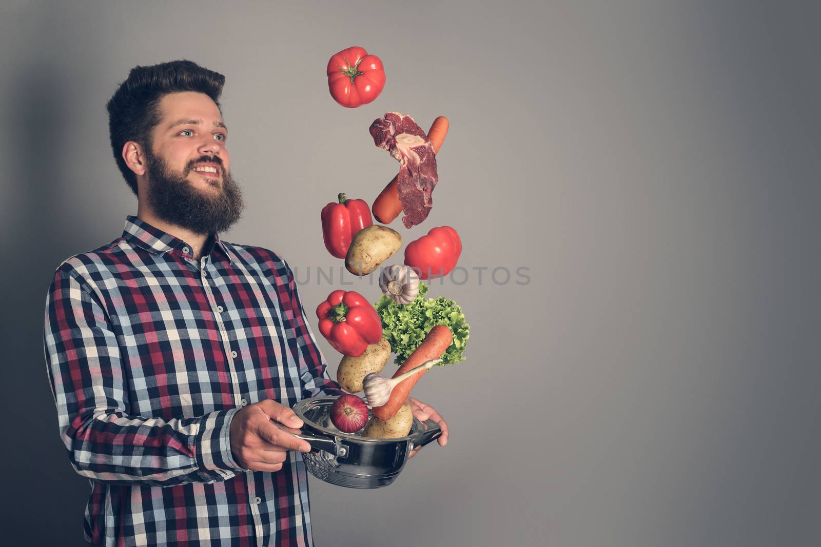 Cooking man concept, smiling bearded man in checked shirt, drop up meat and vegetables from a pan, studio shot on gray background