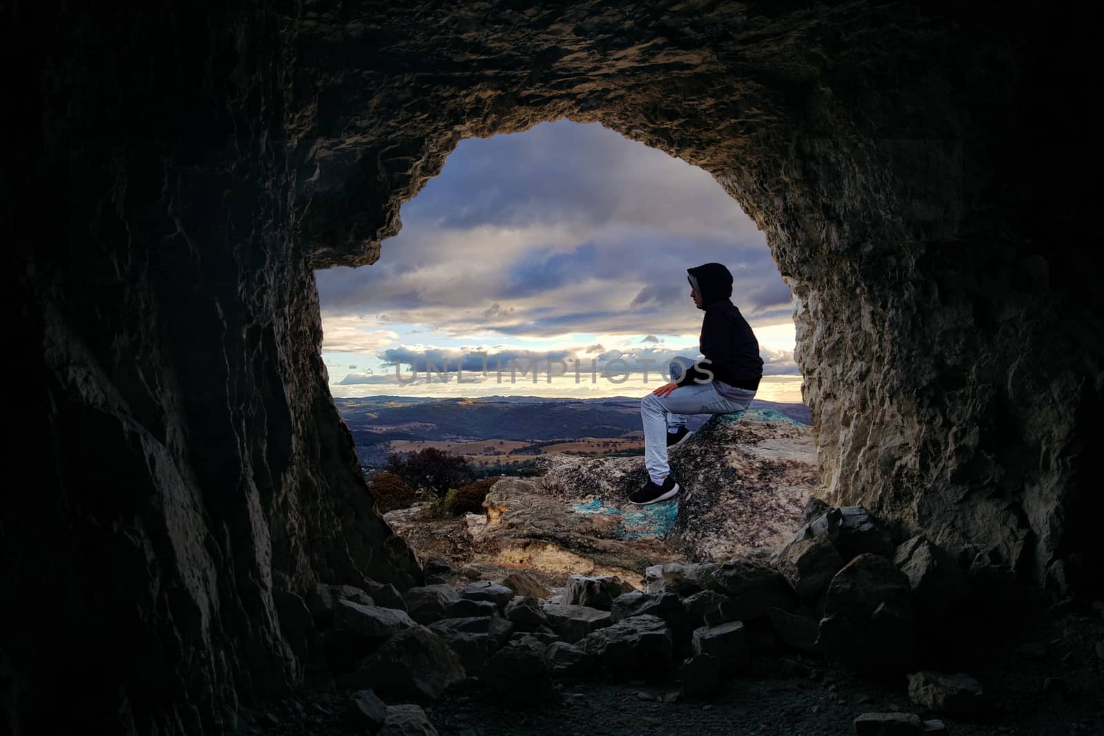 Looking through a cave to a male sitting on a rocky outcrop that looks over the valley