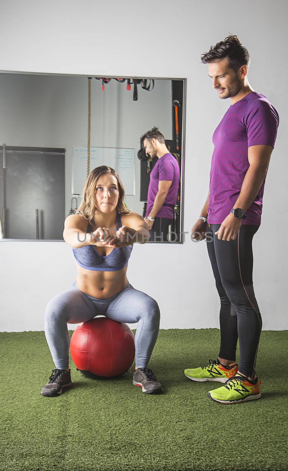 Young woman doing a squat on a red medicine ball with a trainer watching her