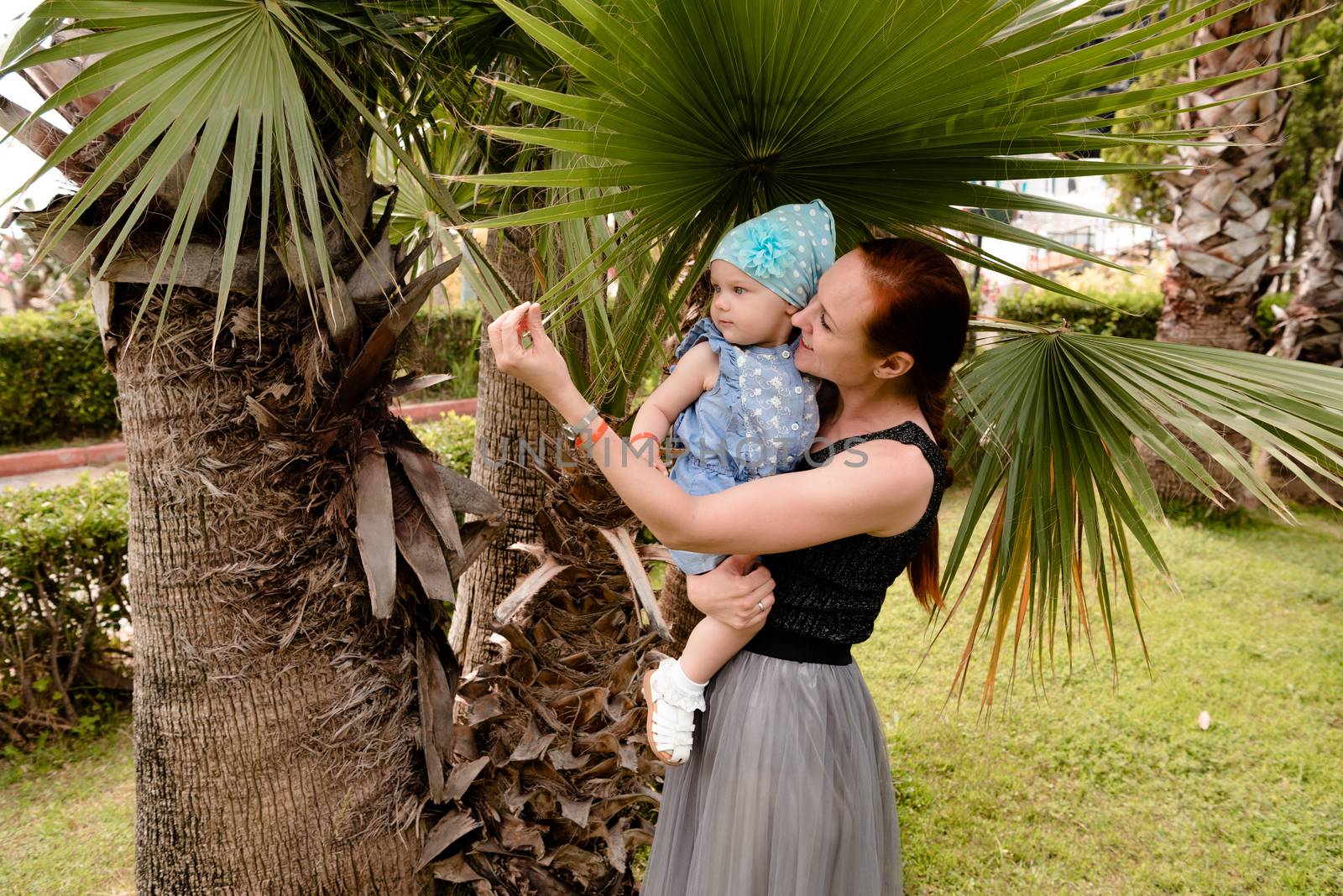 a little girl sits in her mother's arms and examines the leaf of a palm tree. she's wearing a blue suit and a hat. In a tropical Park