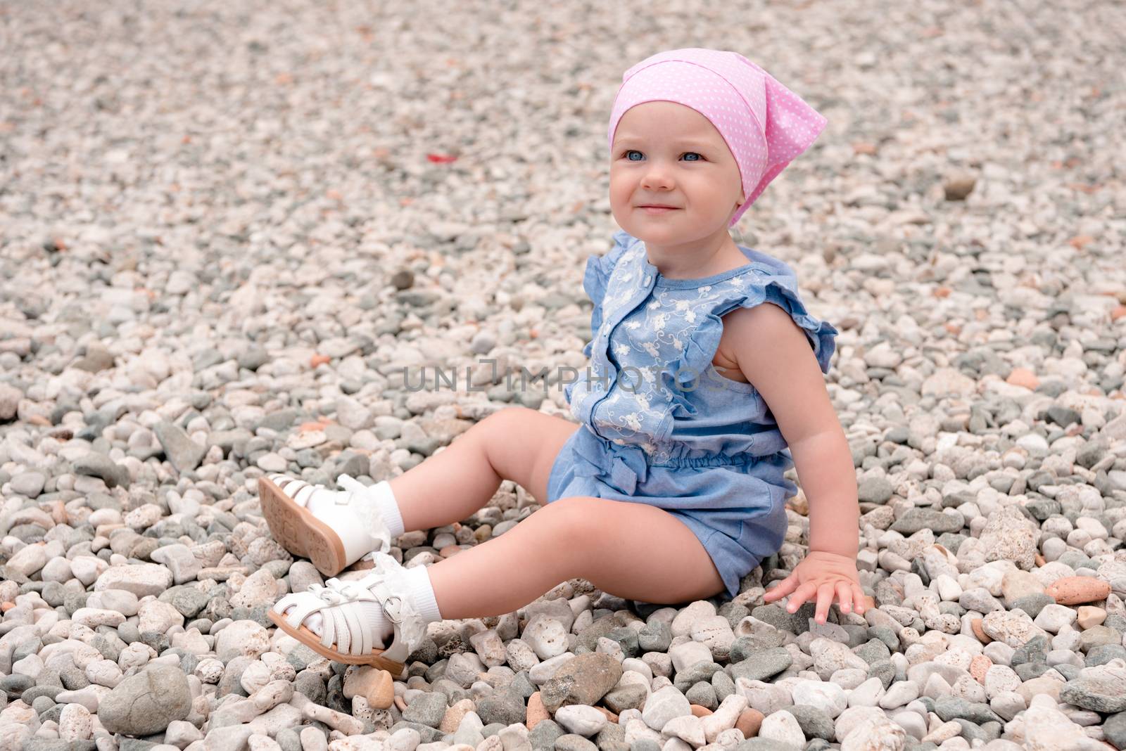 the baby in a pink cap, blue overalls, white socks and sandals sits on a large number of small stones