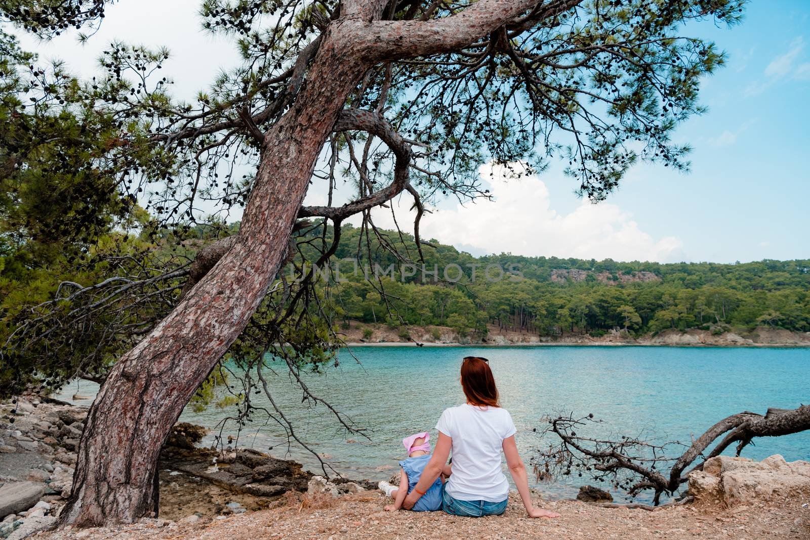 girl and child sitting on a pebble next to a spruce tree h background of blue sea and green hills