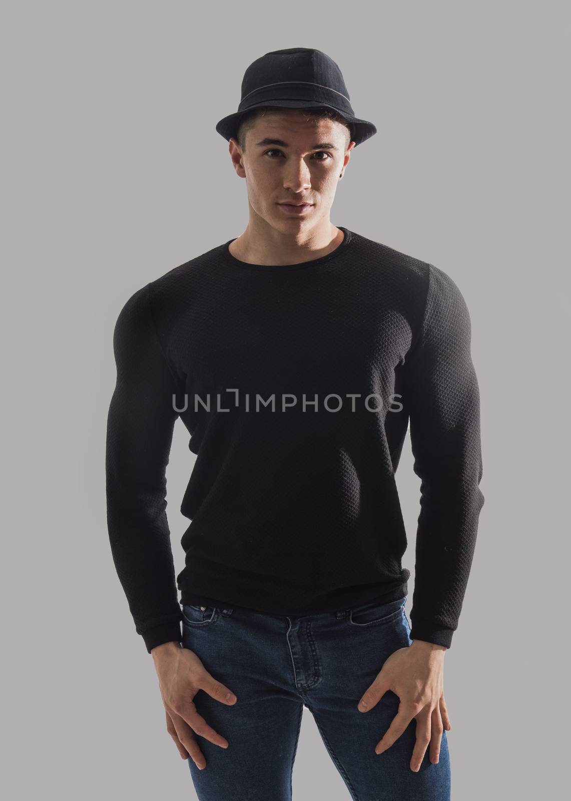 Handsome young muscular man looking at camera in studio shot, wearing fedora hat, with arms crossed on chest, on grey background
