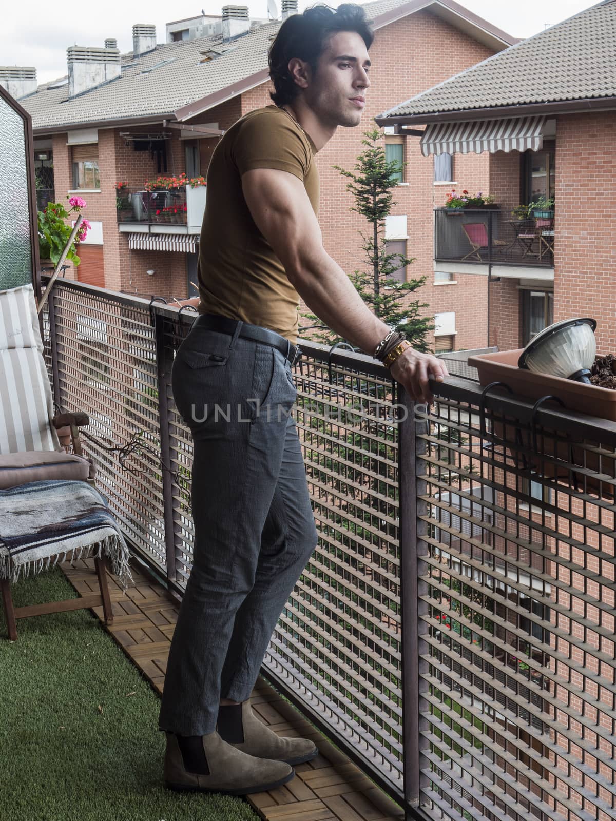 Handsome dark haired young man standing in a balcony, looking out and down at the view, wearing brown T-shirt, in summer, full length shot