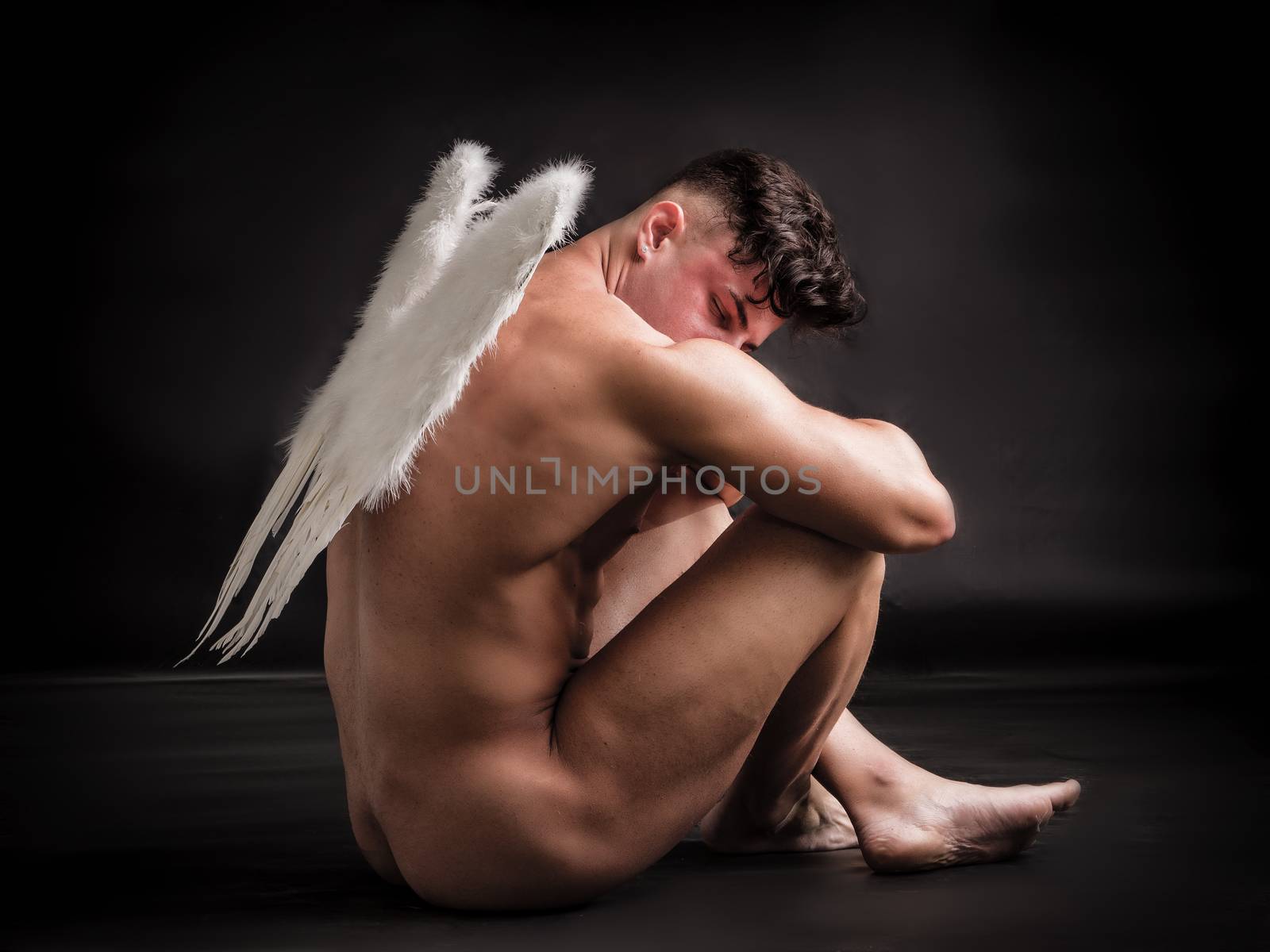Fallen naked male angel sitting in dark with white feather wings spreading from his back. Unrecognizable young man