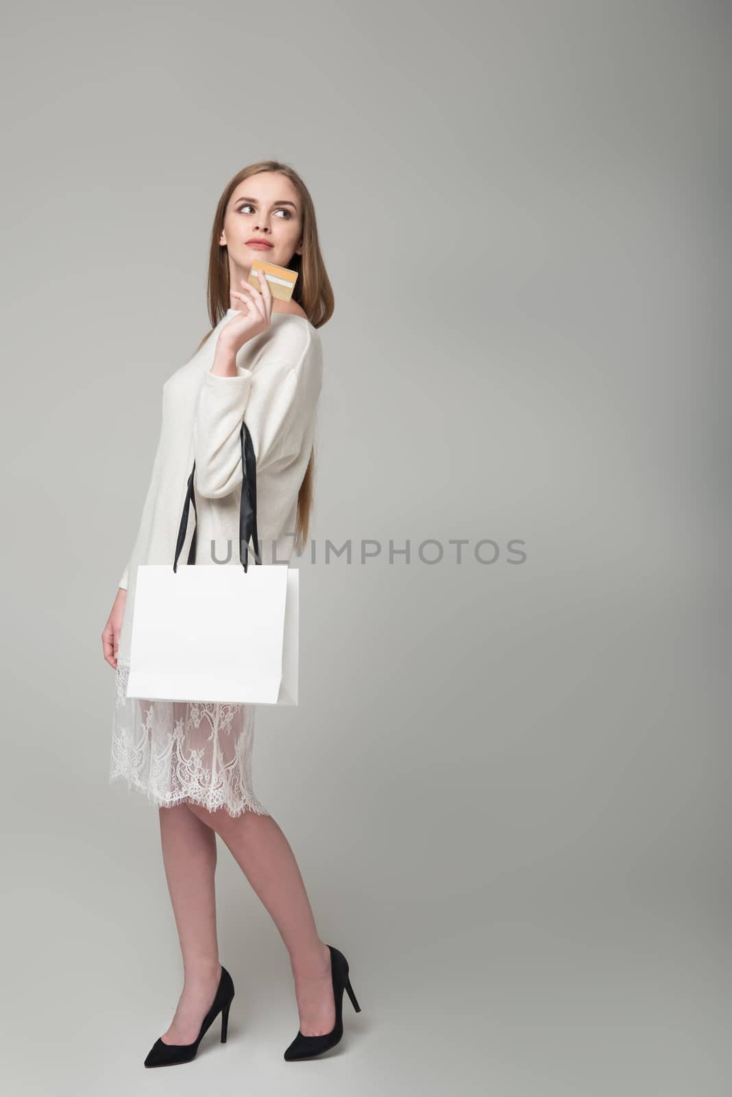 Young model long-haired blond girl in white short dress with lace stands holding white package with copy space