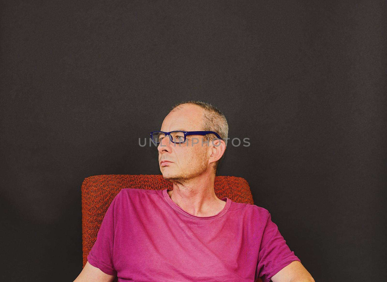 Creative portrait. Middle aged man with glasses sitting in an armchair. Portrait of mature man on black background by roman_nerud