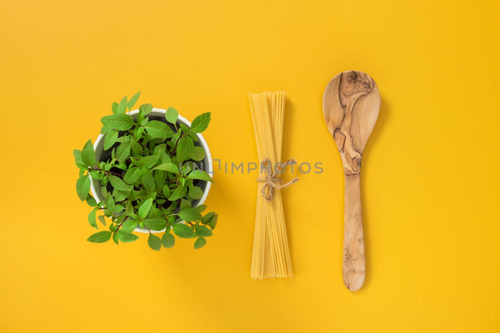 Cooking pasta. Basil herbs in a pot, spaghetti and wooden spoon on yellow background.