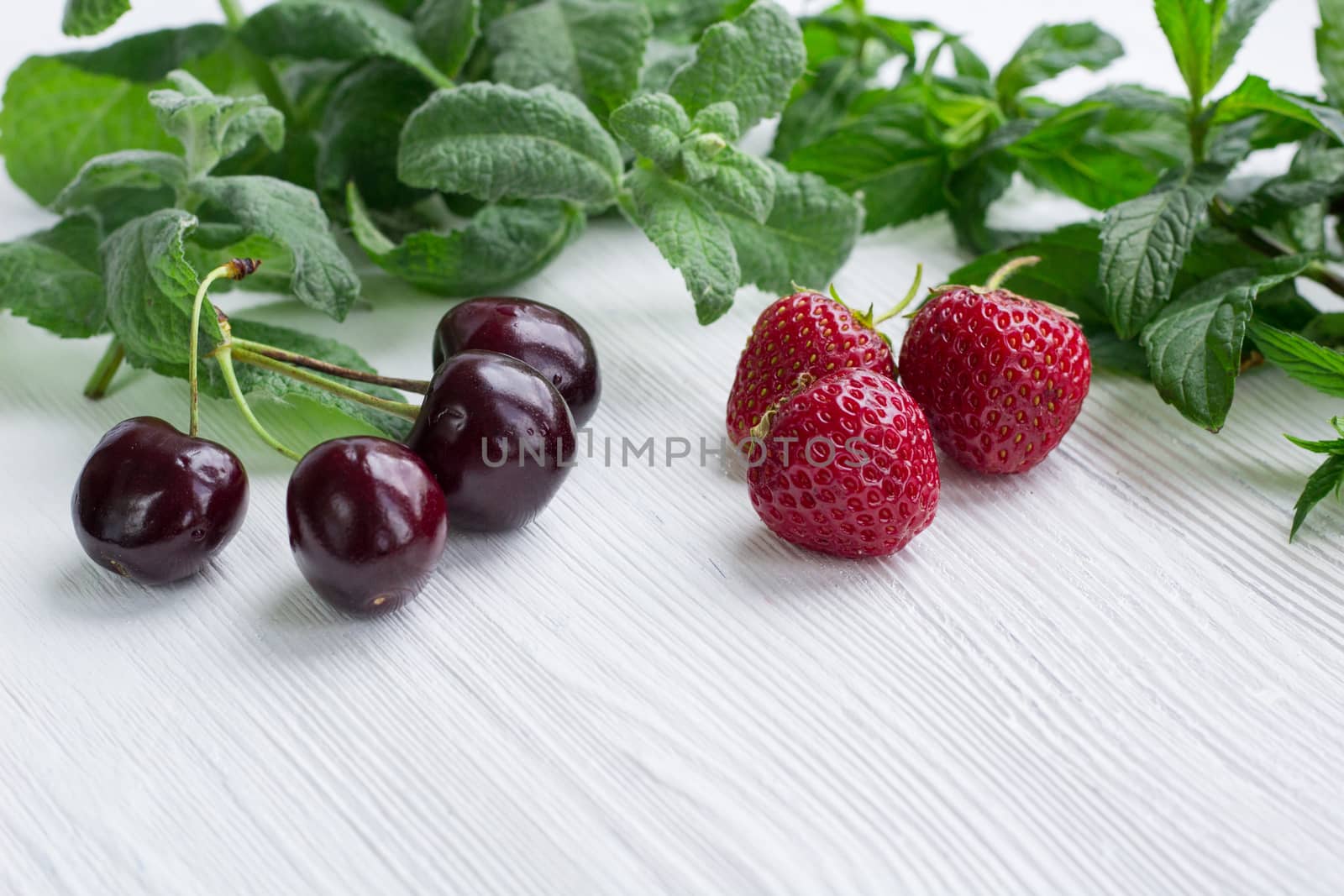 Vinous cherries, red strawberries with green herbal mix of fresh mint and melissa herbs on white wooden background