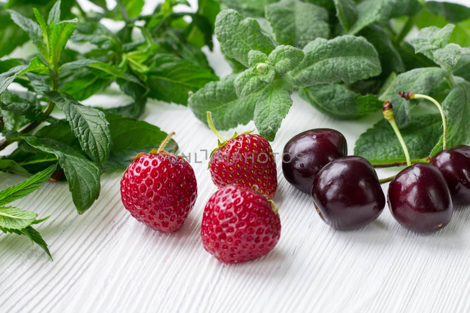 Vinous cherries, red strawberries with green herbal mix of fresh mint and melissa herbs on white wooden background