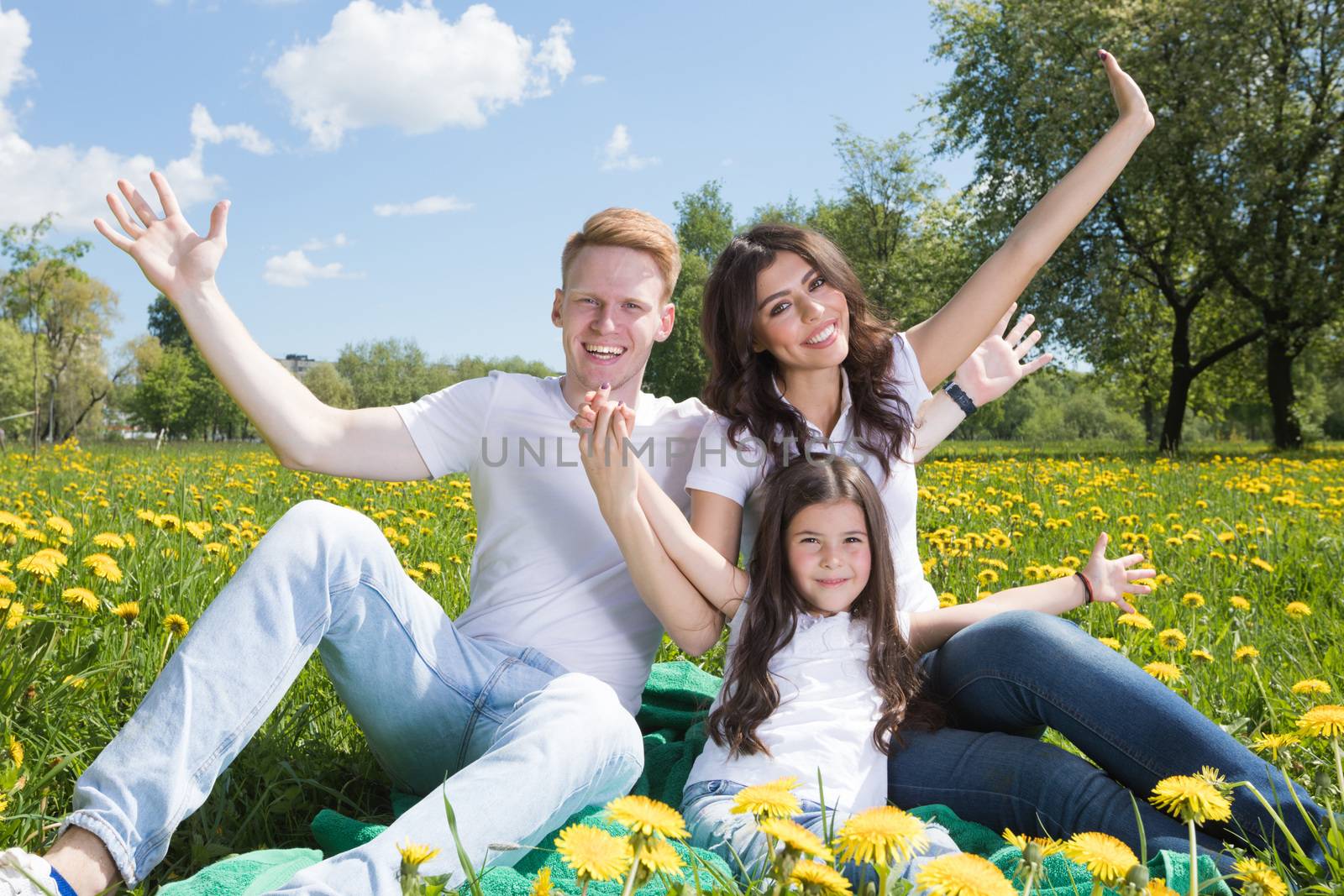 Portrait of happy smiling family of parents and girl sitting on grass with dandelion flowers at sunny summer day