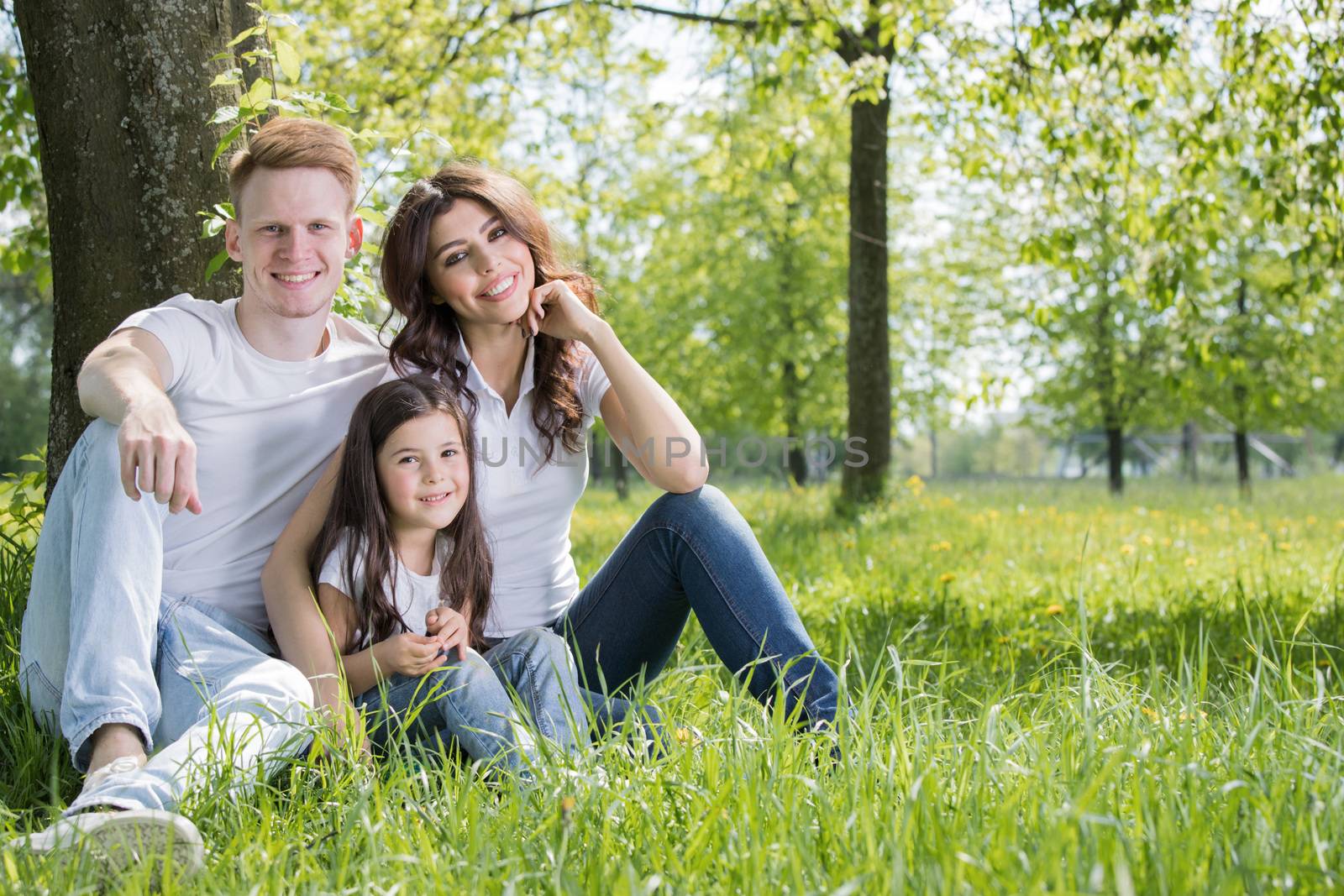 Portrait of happy family sitting on grass under a tree in park on a sunny day