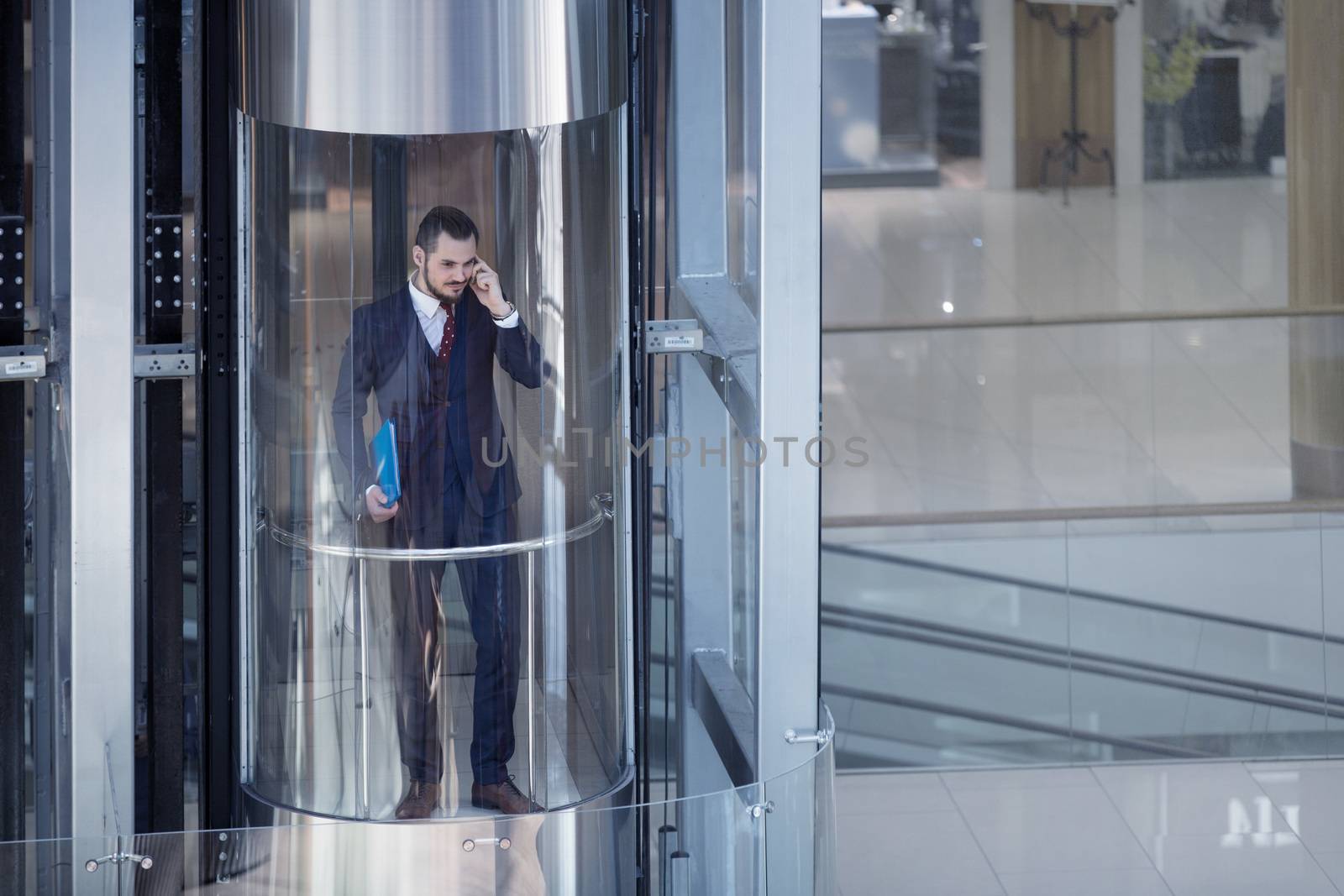 Businessman in modern glass elevator talking by the phone and holding papers