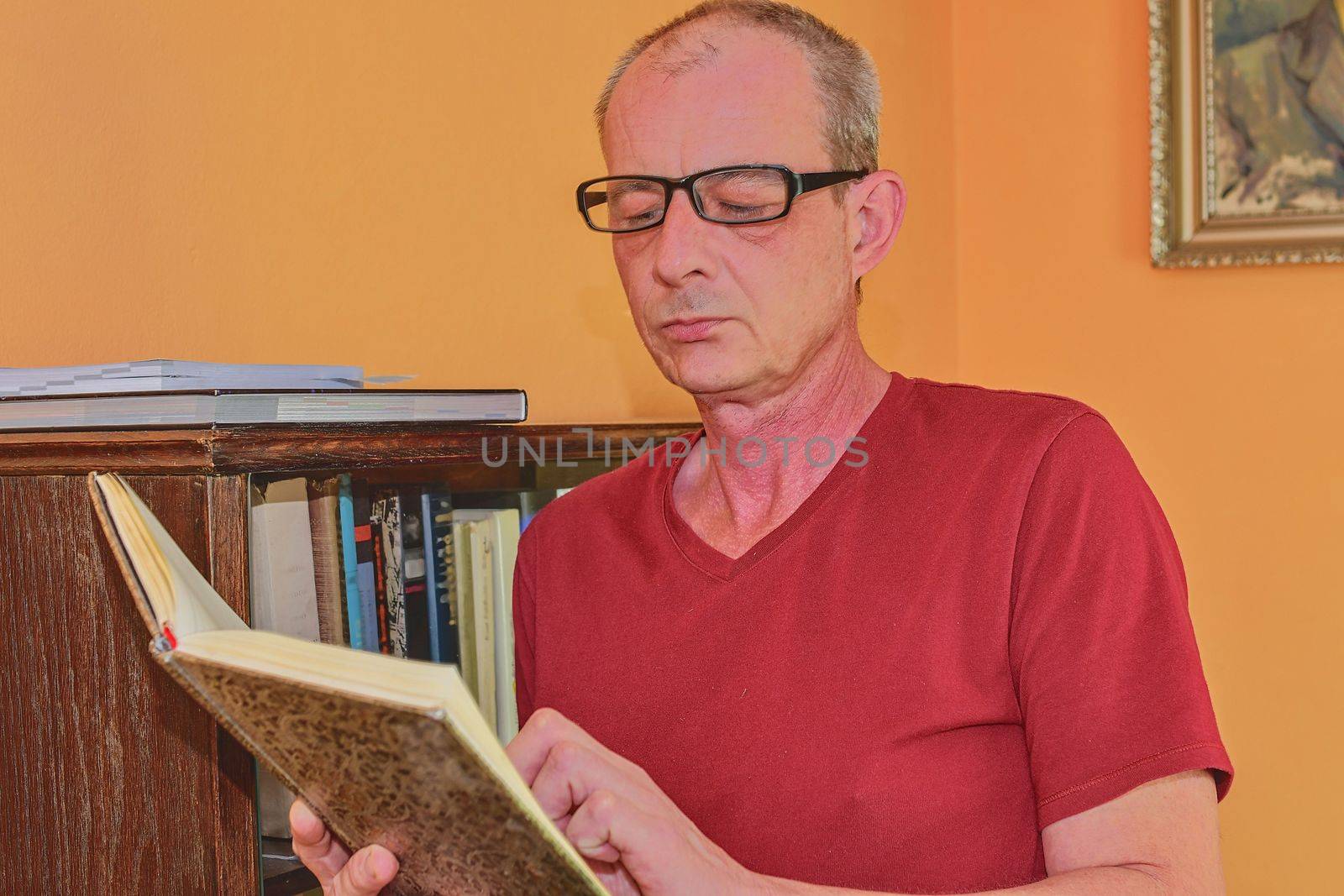 Middle aged man man is reading book in living room. Mature man is standing next to bookcase. 