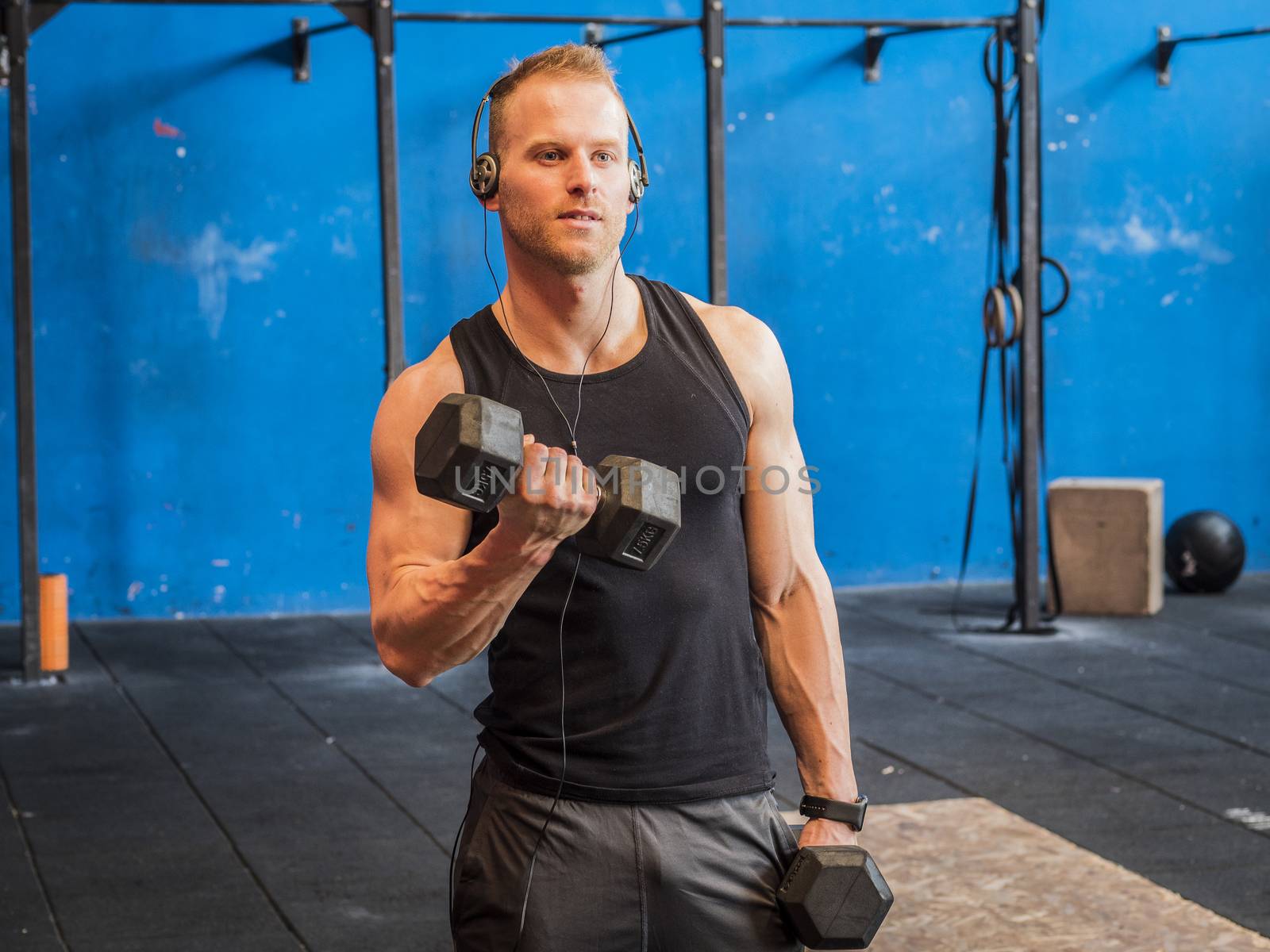 Handsome blond young man training biceps lifting dumbbells, standing in a gym, looking to a side