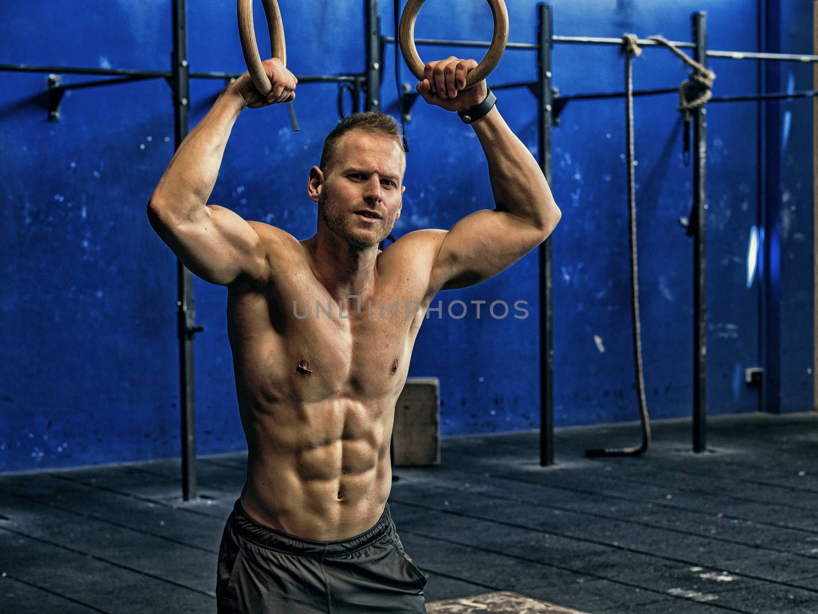 Handsome shirtless muscular young man athlete exercising in gym, hanging rings.