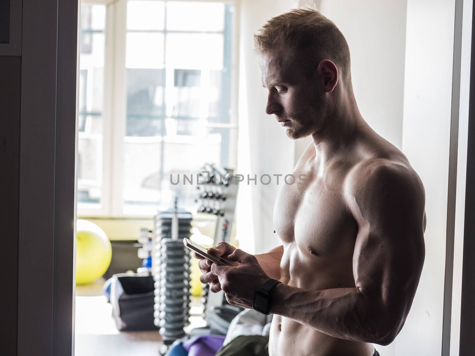 Shirtless muscular blond young man using cell phone in gym