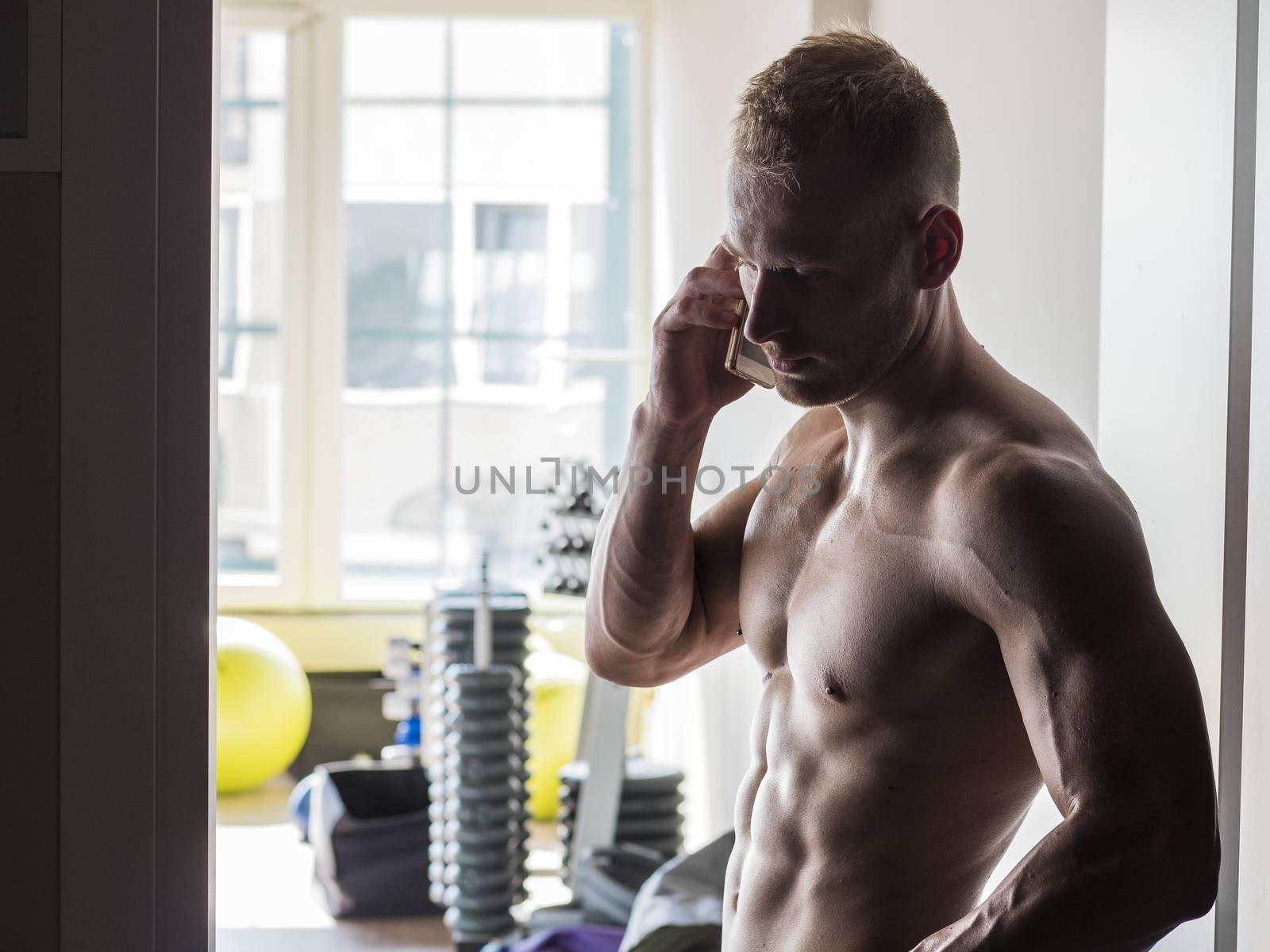 Shirtless muscular blond young man using cell phone in gym, calling somebody