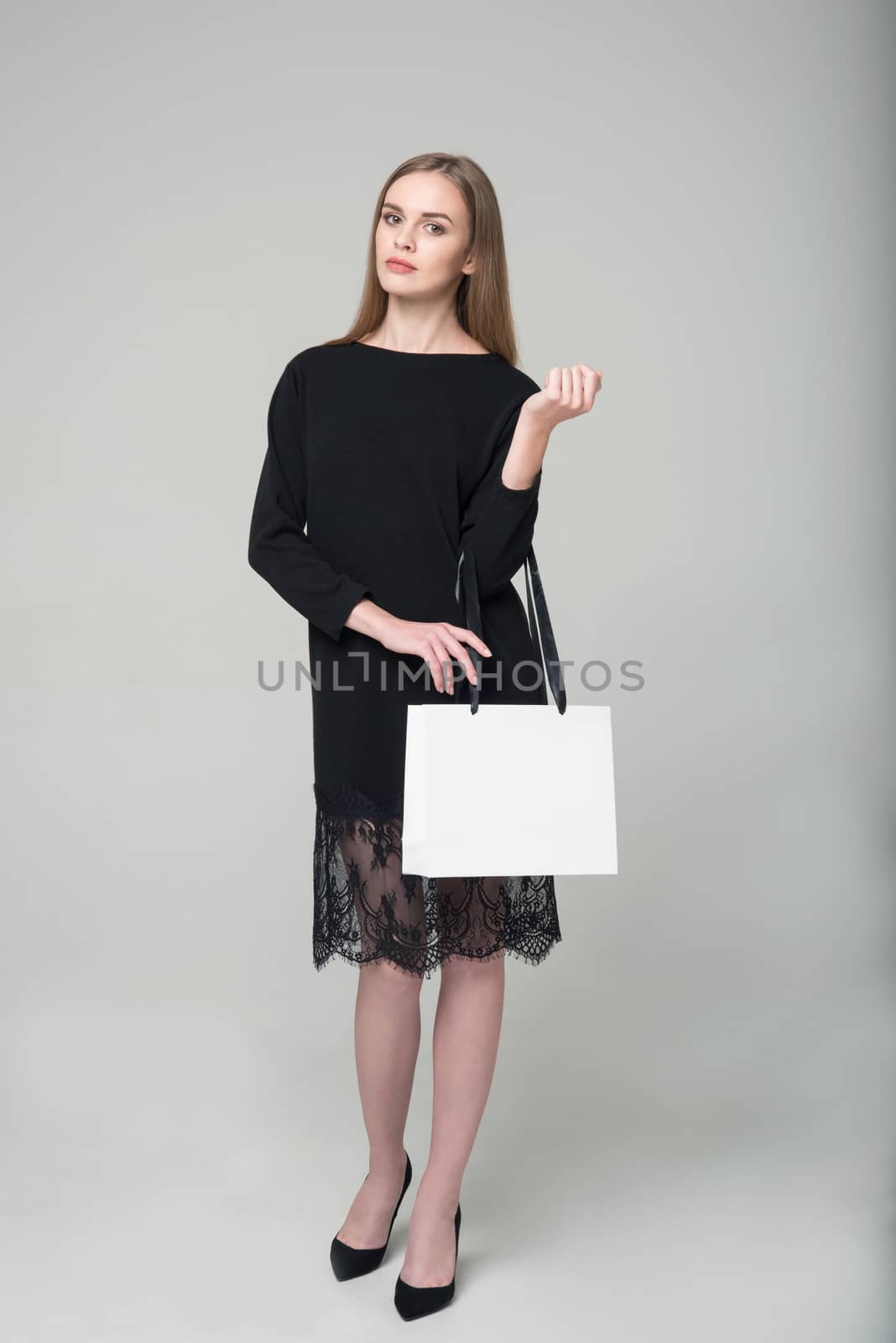 Young model long-haired blond girl in black short dress with lace stands holding white package with copy space