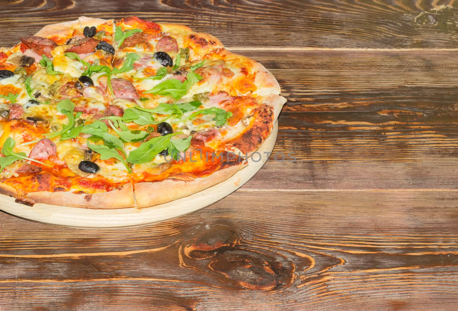 Fragment of the cooked round pizza with sausages, mushrooms, olives and arugula on the old wooden table
