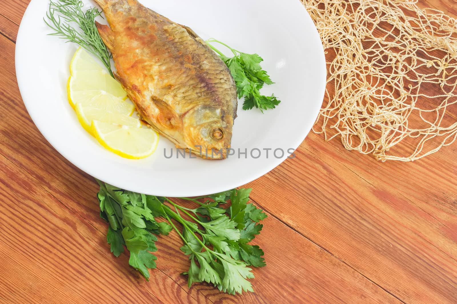 Fragment of the white dish with fried crucian, slices of the lemon, twigs od the parsley and dill and part of the fishing net on a surface of the old wooden planks
