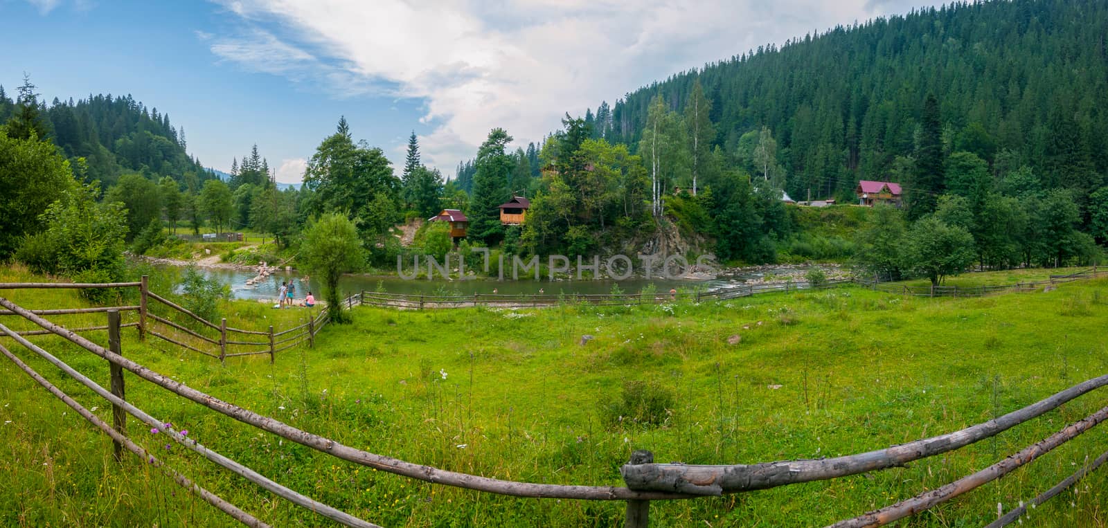 Beautiful views of the mountainous terrain with the current river between the low banks with green grass on one side and trees and houses on the other. by Adamchuk