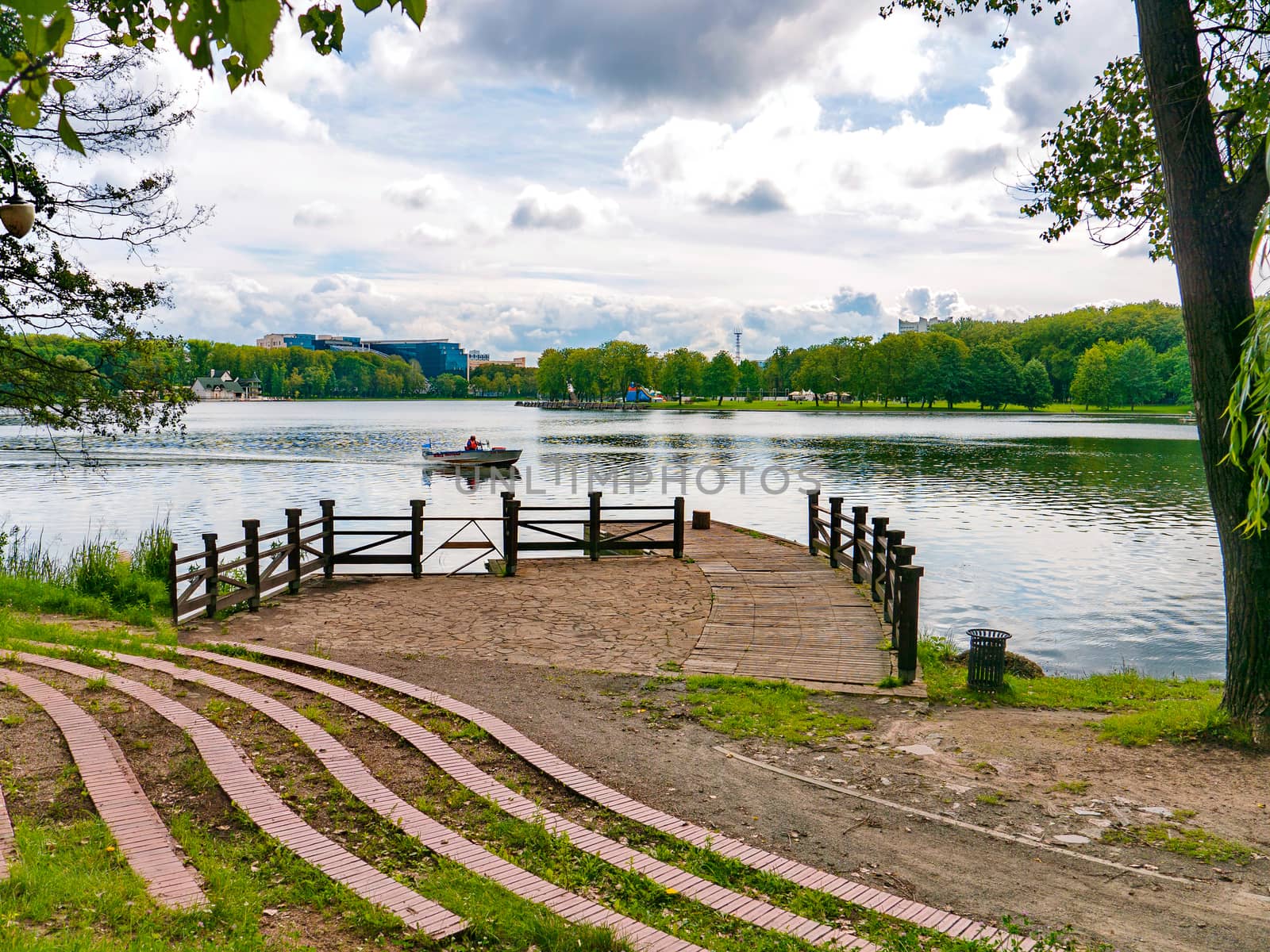 a path to the pier on the lake with a boat, a cloudy sky and green trees around. resting place, fishing